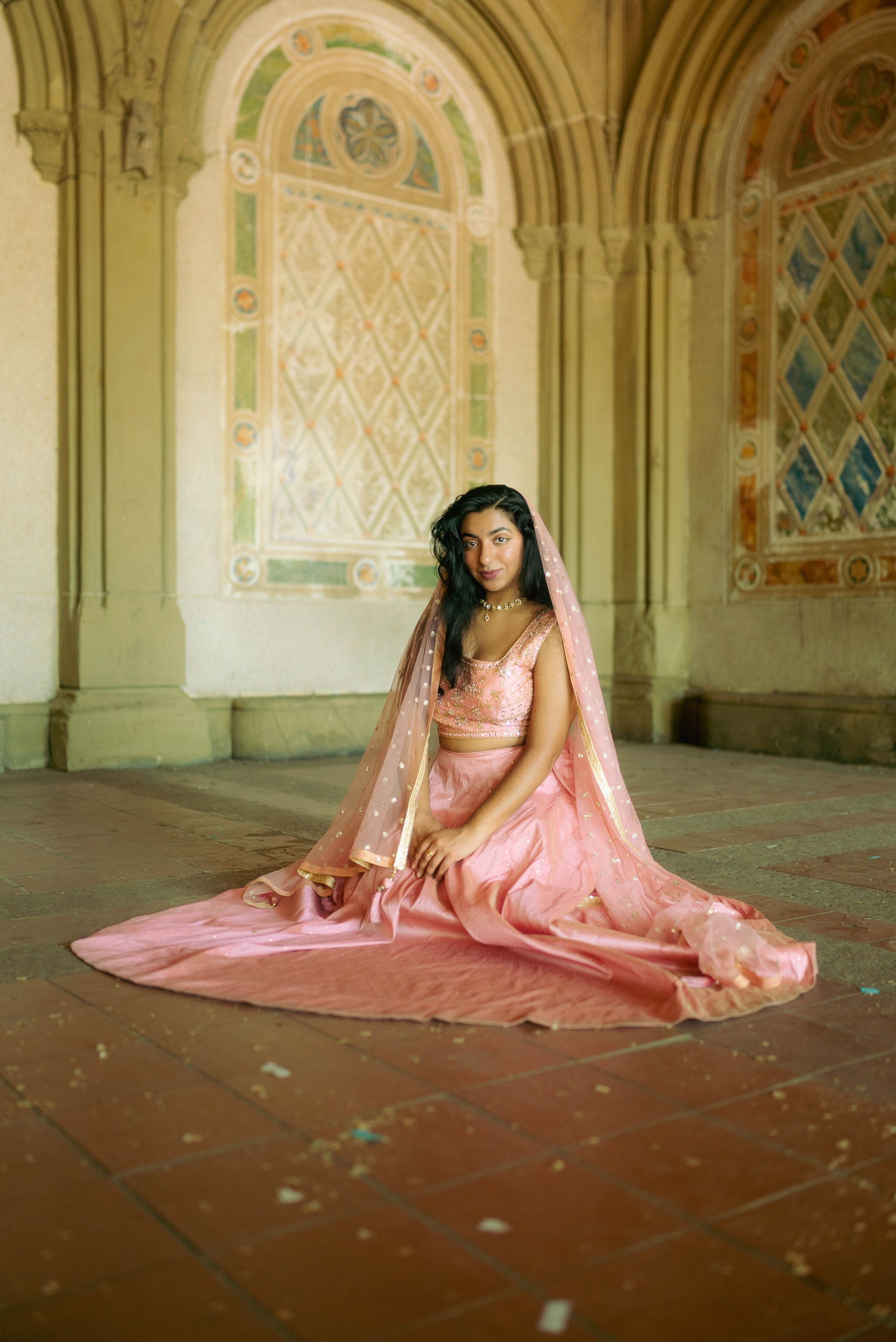 A person in a flowing pink traditional dress sits gracefully in an ornate architectural space with arched windows.