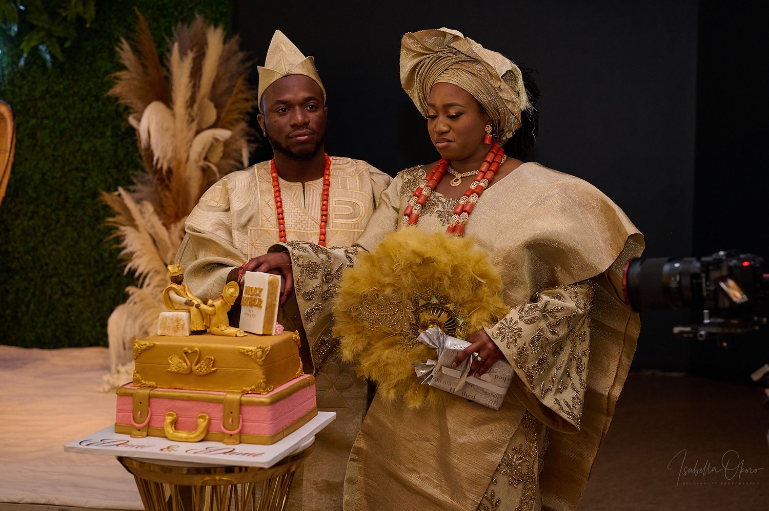 A couple in traditional Nigerian attire stands beside a decorative wedding cake.