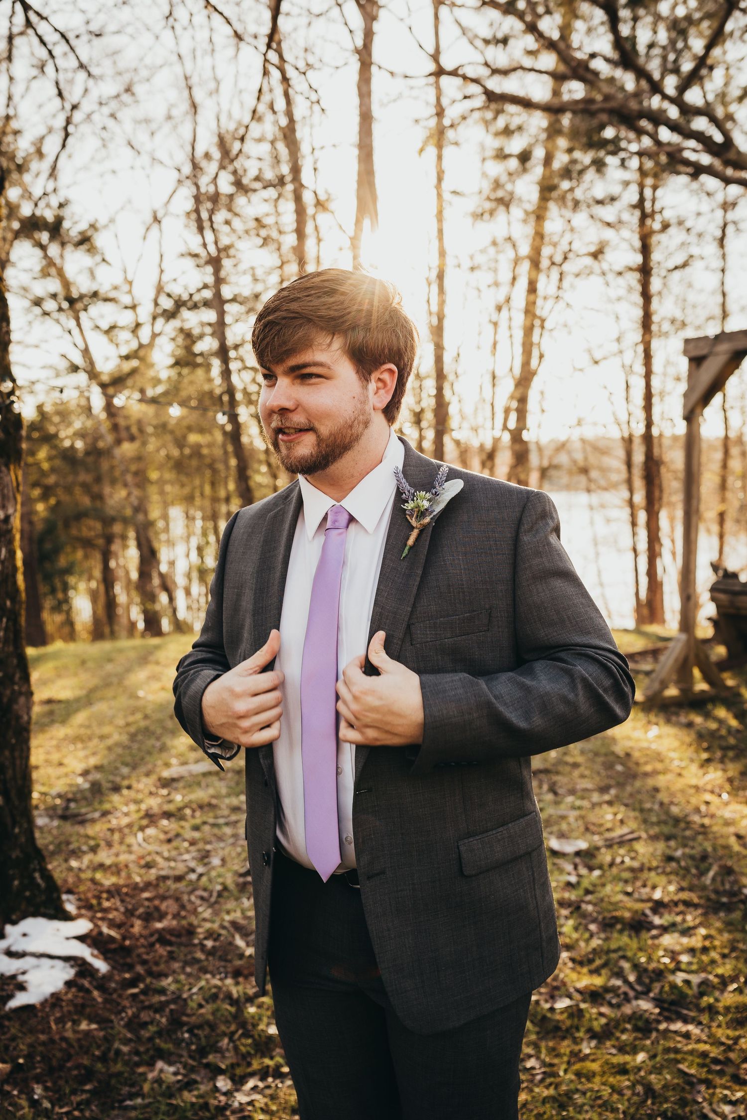 A person in a dark suit and lavender tie adjusts their jacket in an autumn forest setting.