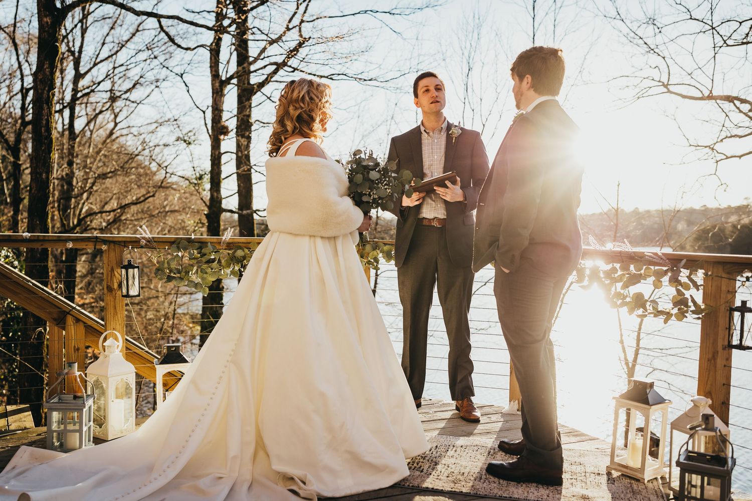 Sunset wedding ceremony on a waterfront deck with bare winter trees in background.