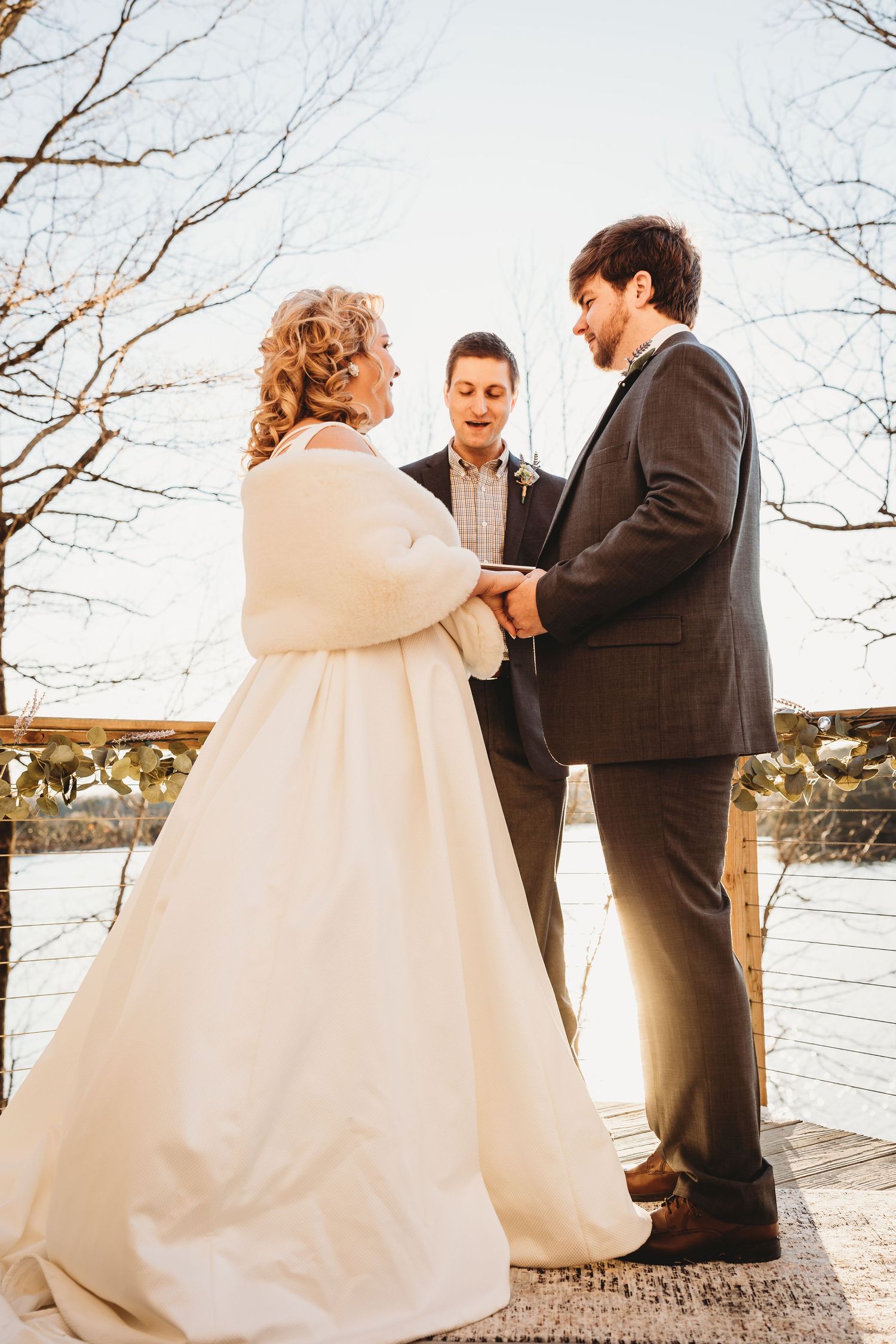 A couple shares an intimate moment during their winter wedding ceremony on a wooden deck overlooking a lake.
