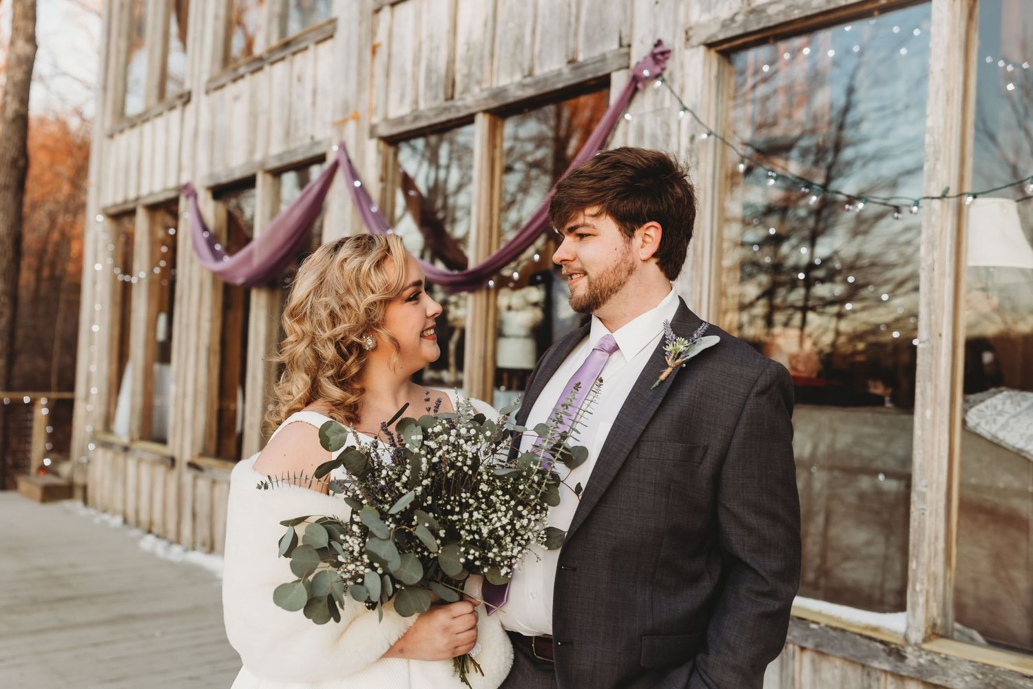 Rustic winter wedding couple poses together against weathered wooden barn exterior.