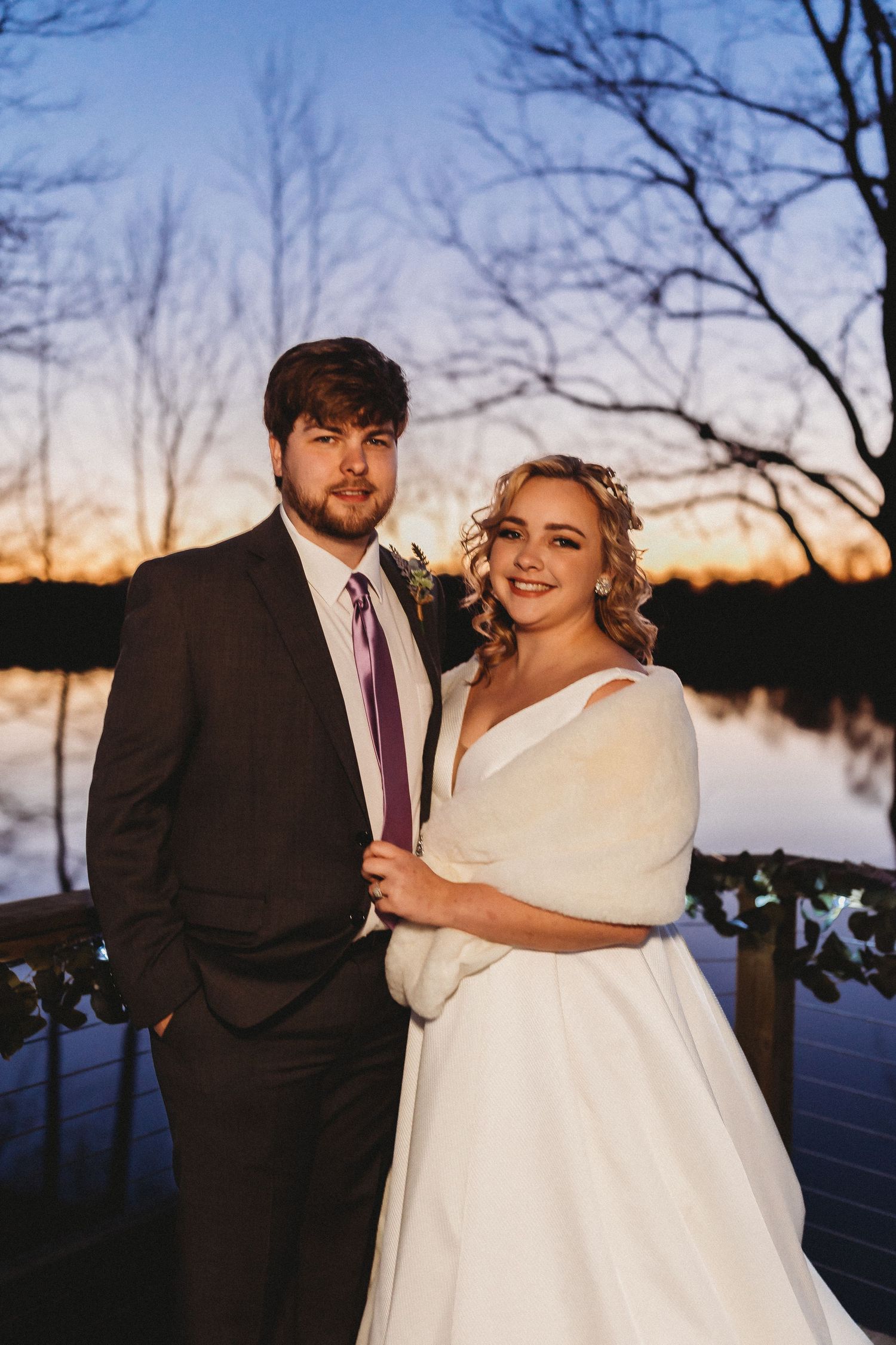 Winter wedding couple poses at sunset near a lake with bare trees in background.