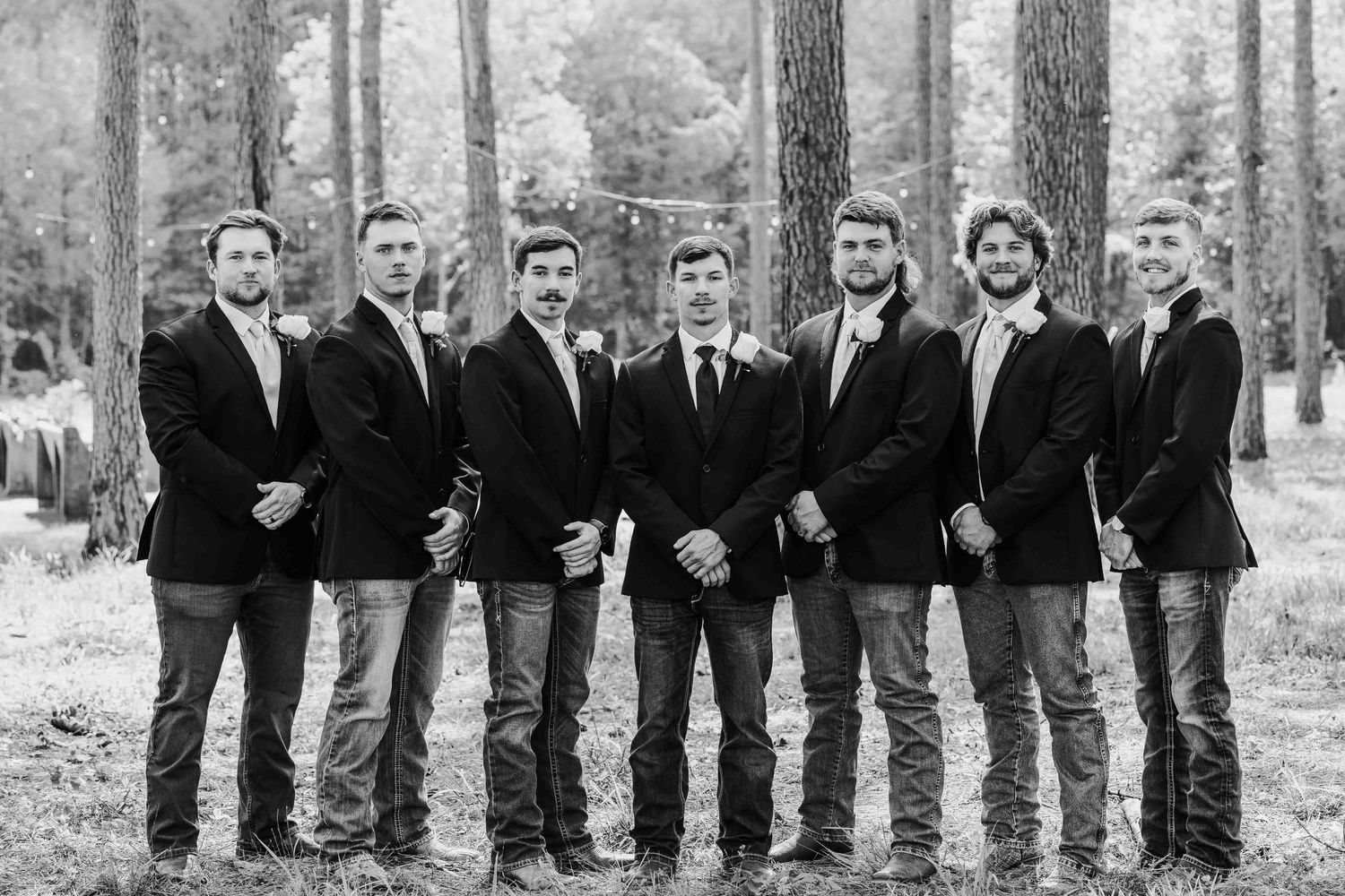 Groomsmen in dark suits pose together in wooded forest setting.