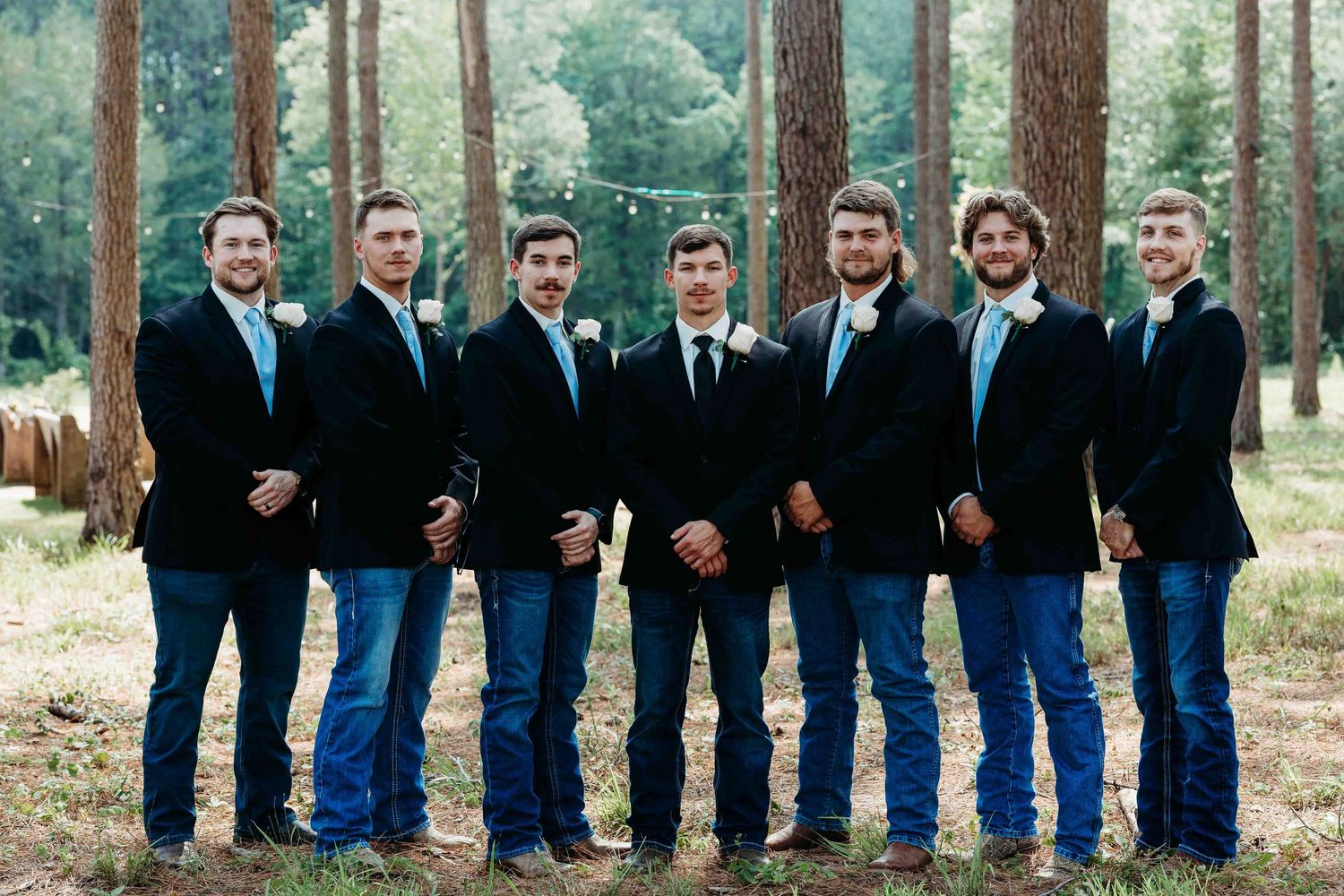 Wedding party members in navy and black attire lined up for photo in pine forest.