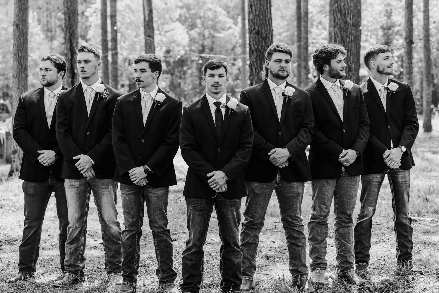 Groomsmen lined up in black suits and jeans pose in a forest setting for a wedding photo.