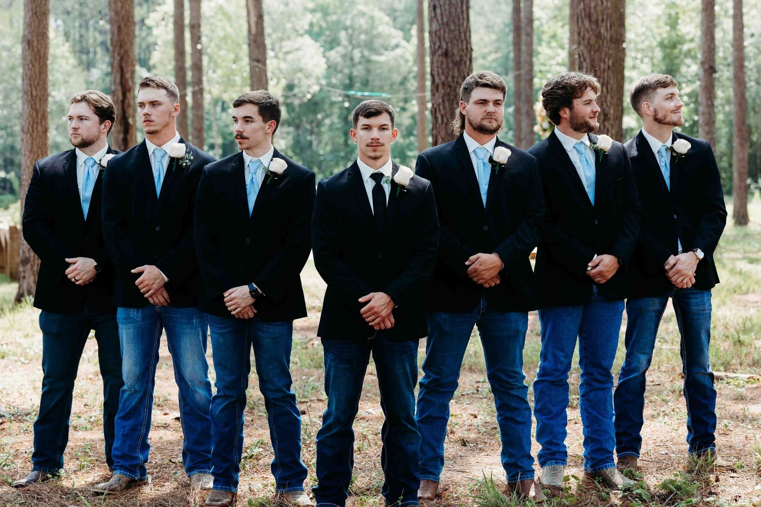 Row of groomsmen in matching dark jackets and blue jeans stand among tall pine trees.