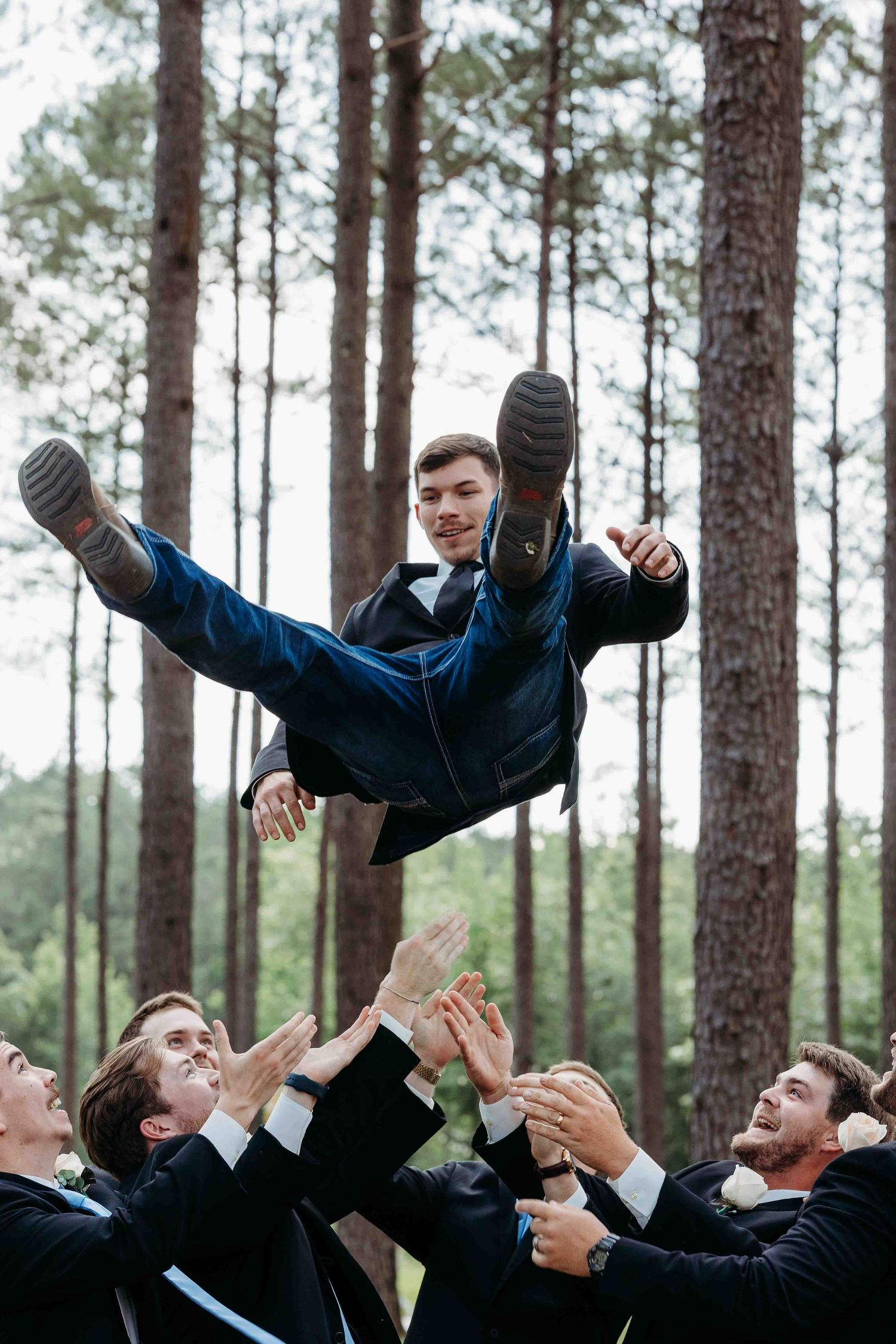 Groomsmen toss someone in the air during an outdoor wedding photoshoot in a pine forest.