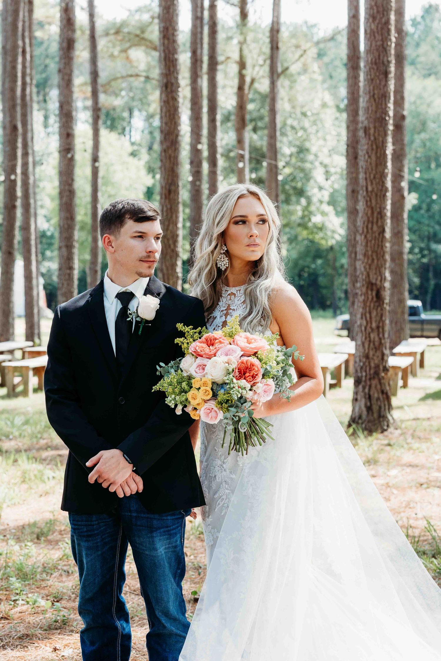A wedding couple stands together in a wooded area with sunlight filtering through tall pine trees.