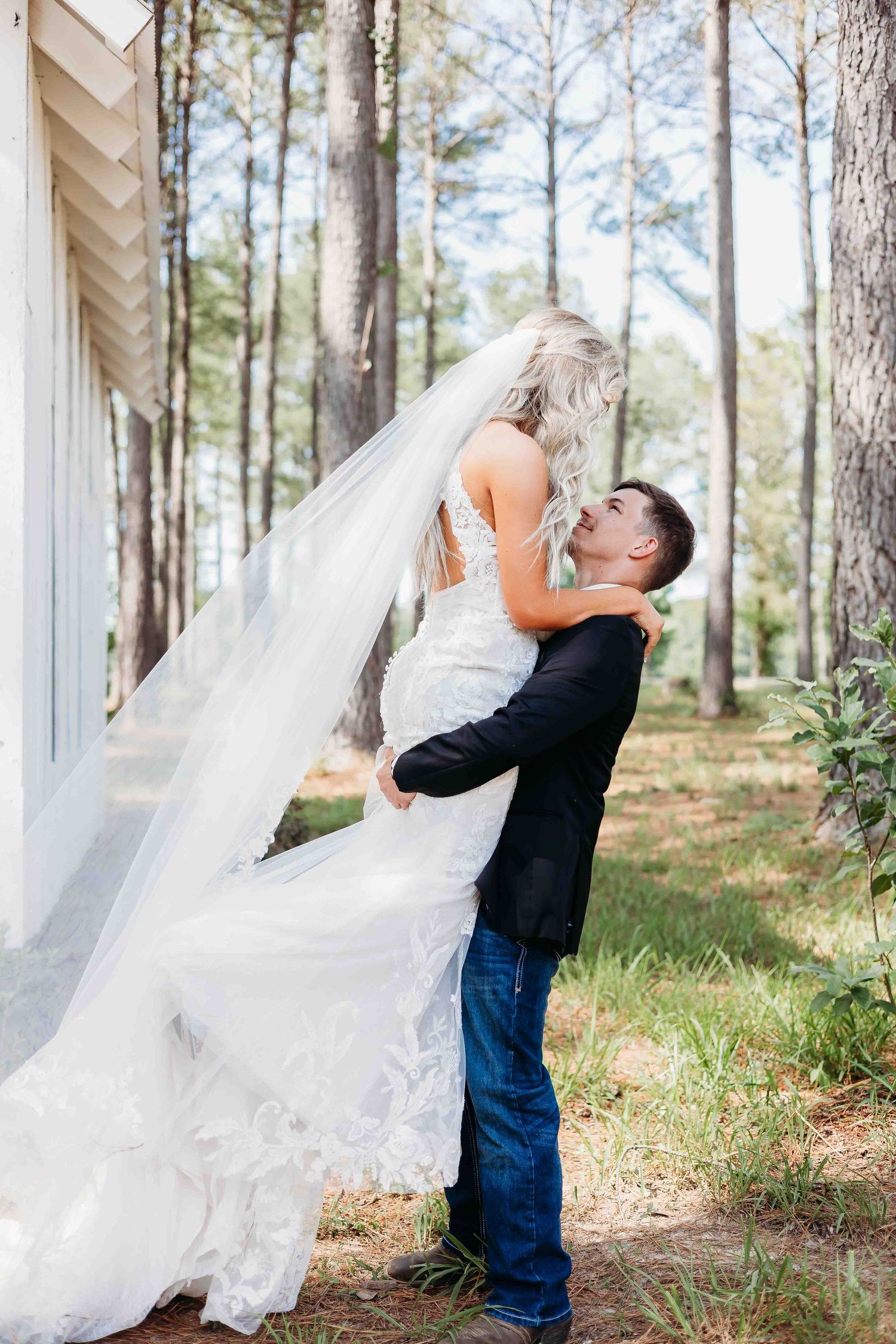 Playful wedding photo of groom lifting bride outside rustic white building among pine trees.