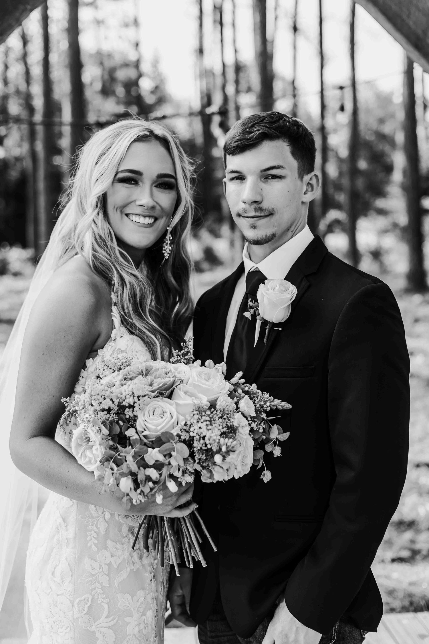 Wedding couple shares intimate moments in a pine forest setting, captured in black and white photography.