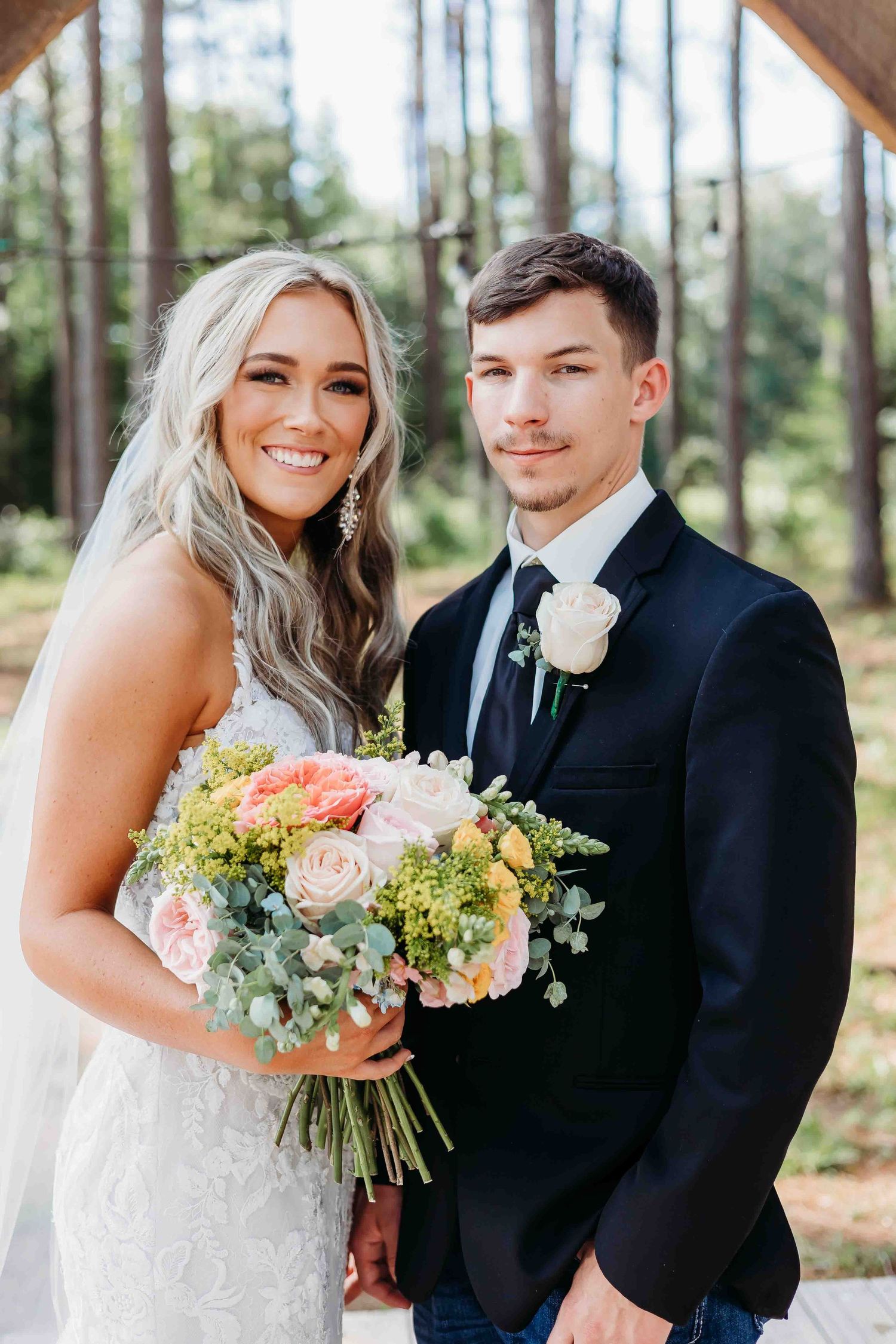 Bride and groom pose together under tall pine trees with colorful floral bouquet.