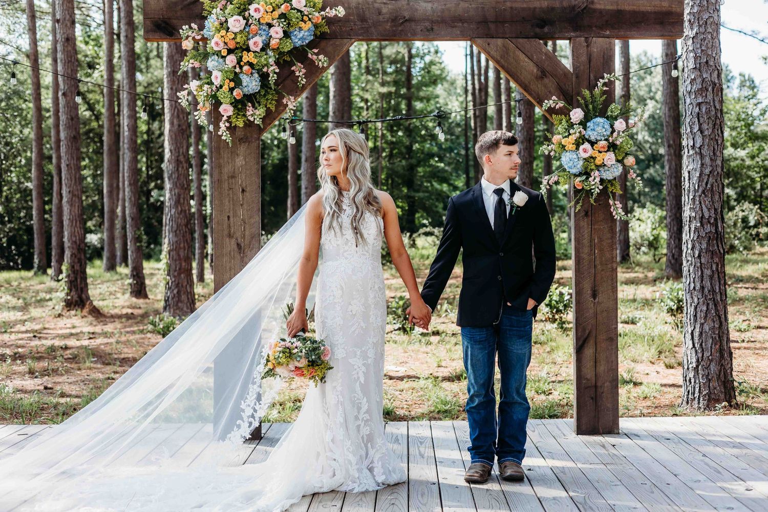 Rustic outdoor wedding ceremony setup with wooden arch decorated with colorful flowers in pine forest.