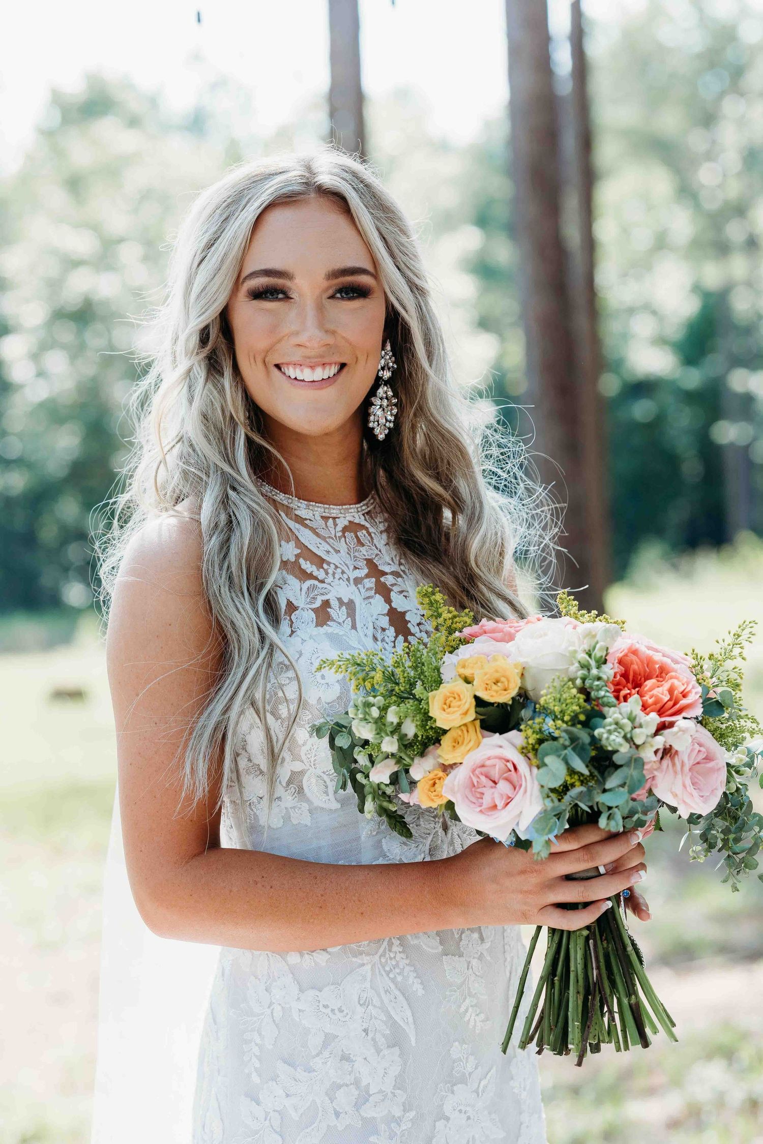 Bride in lace dress holds colorful spring bouquet in sunlit forest setting.