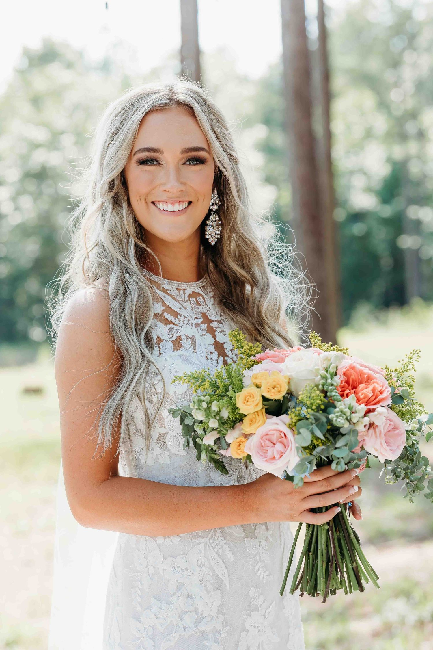 Bride wearing detailed lace gown holds fresh bouquet of coral and yellow flowers in woodland setting.