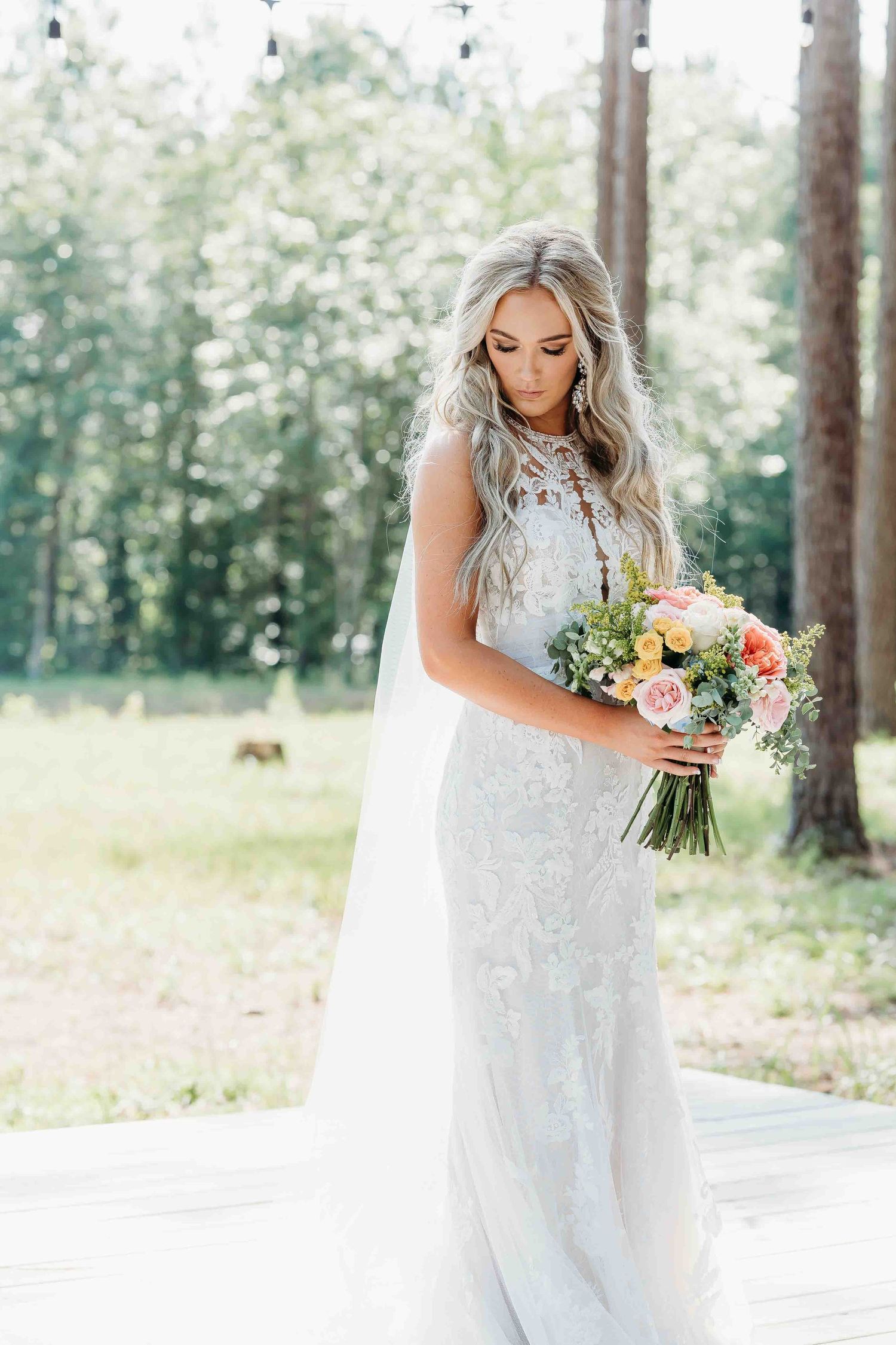 Elegant bride in white lace gown poses with pastel floral bouquet in pine forest.