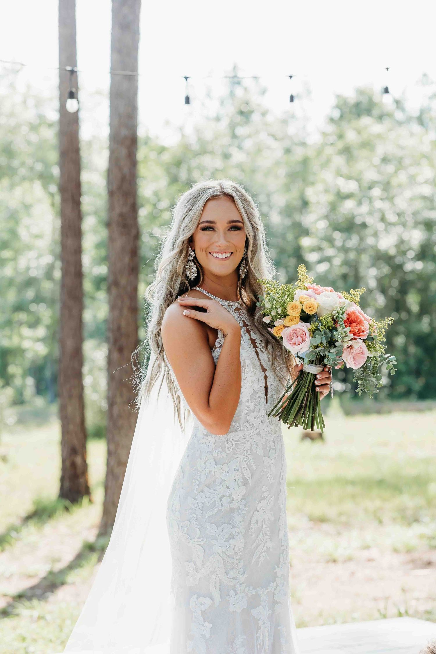A bride in a lace wedding dress poses outdoors among trees holding a colorful bouquet.