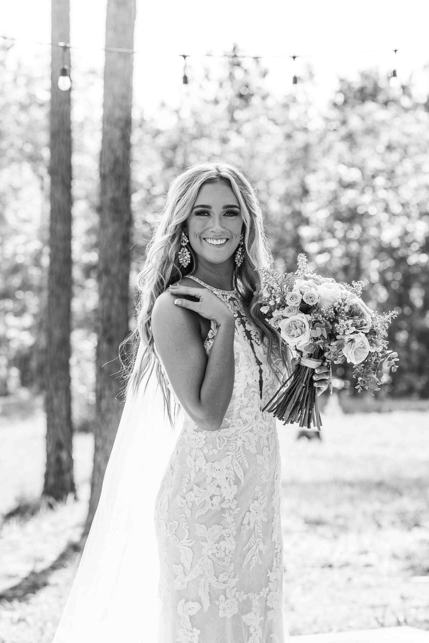 A black and white photo of a bride in a detailed lace gown holding a floral bouquet in a wooded setting.
