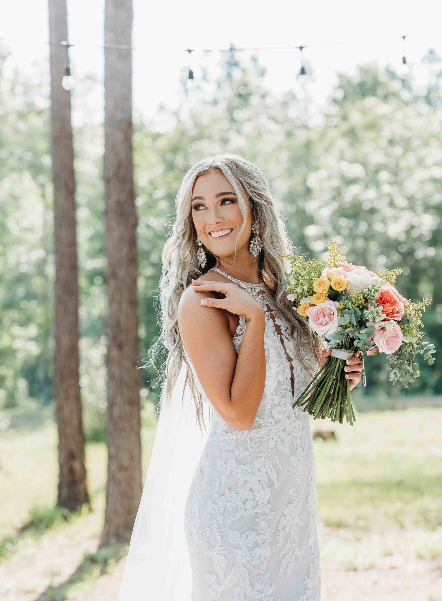 A bride laughs while holding her wedding bouquet in a sun-dappled forest setting.