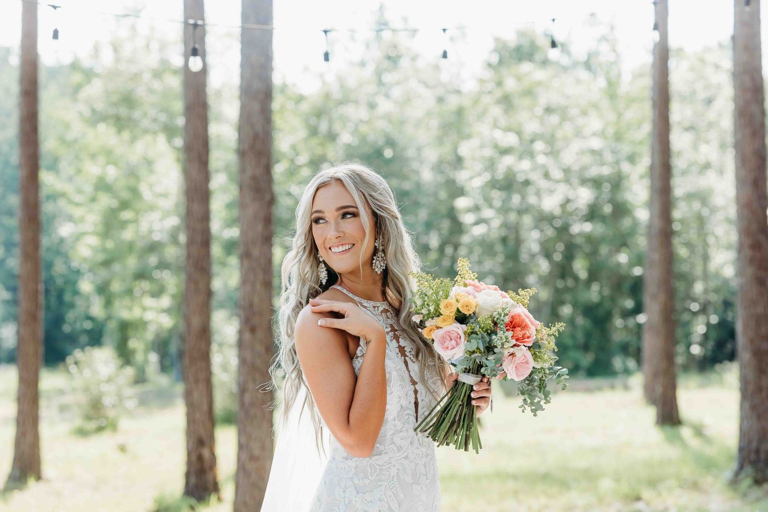 A bride poses with her bouquet in an outdoor forest setting with sunlight filtering through trees.