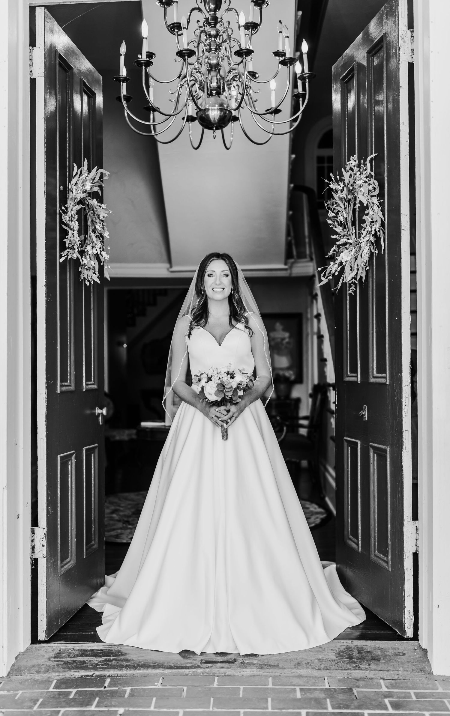 Elegant bride in a flowing white ball gown stands in a doorway beneath a crystal chandelier.