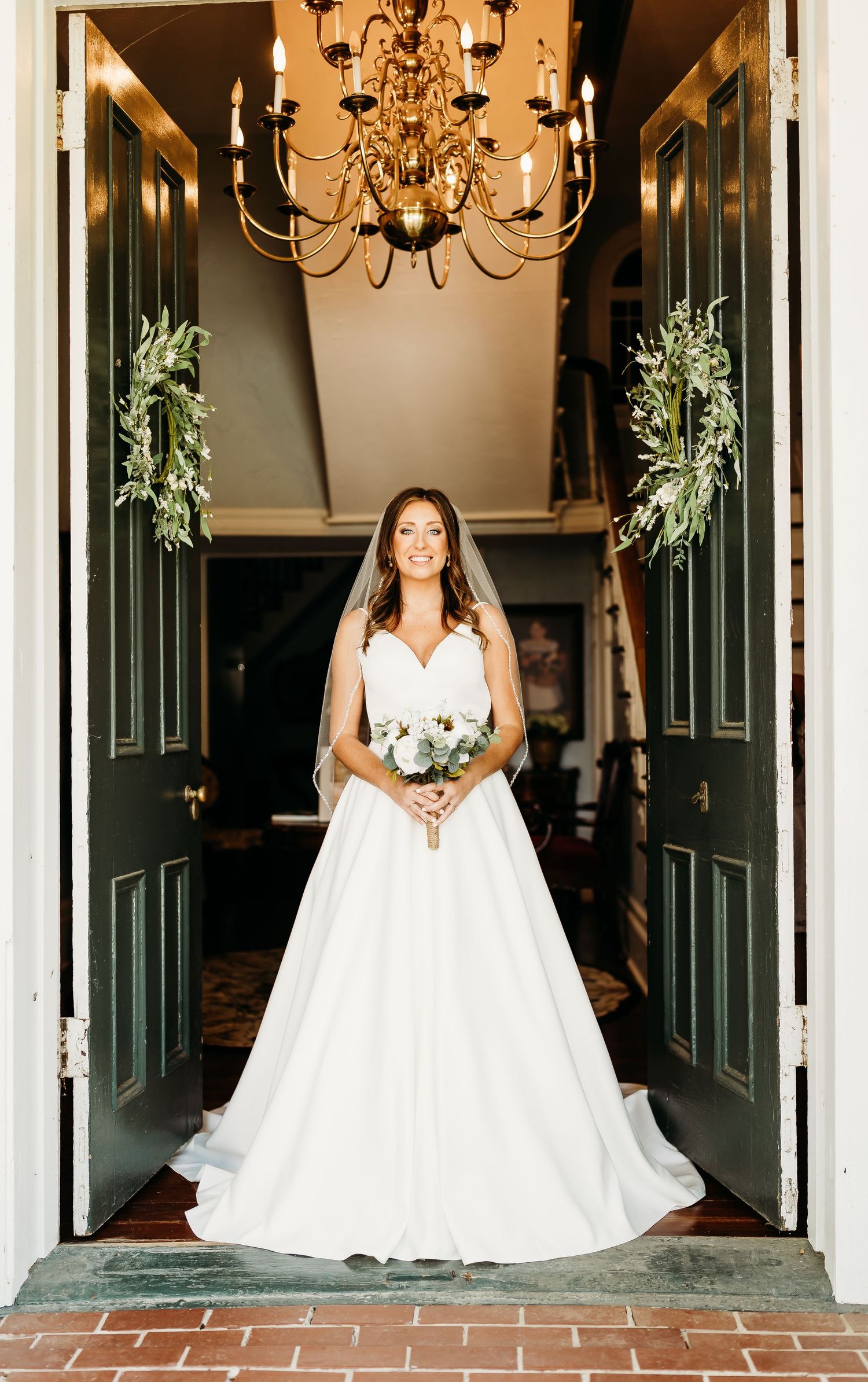 Bride wearing a classic white wedding gown poses in an elegant hallway with green floral decor.