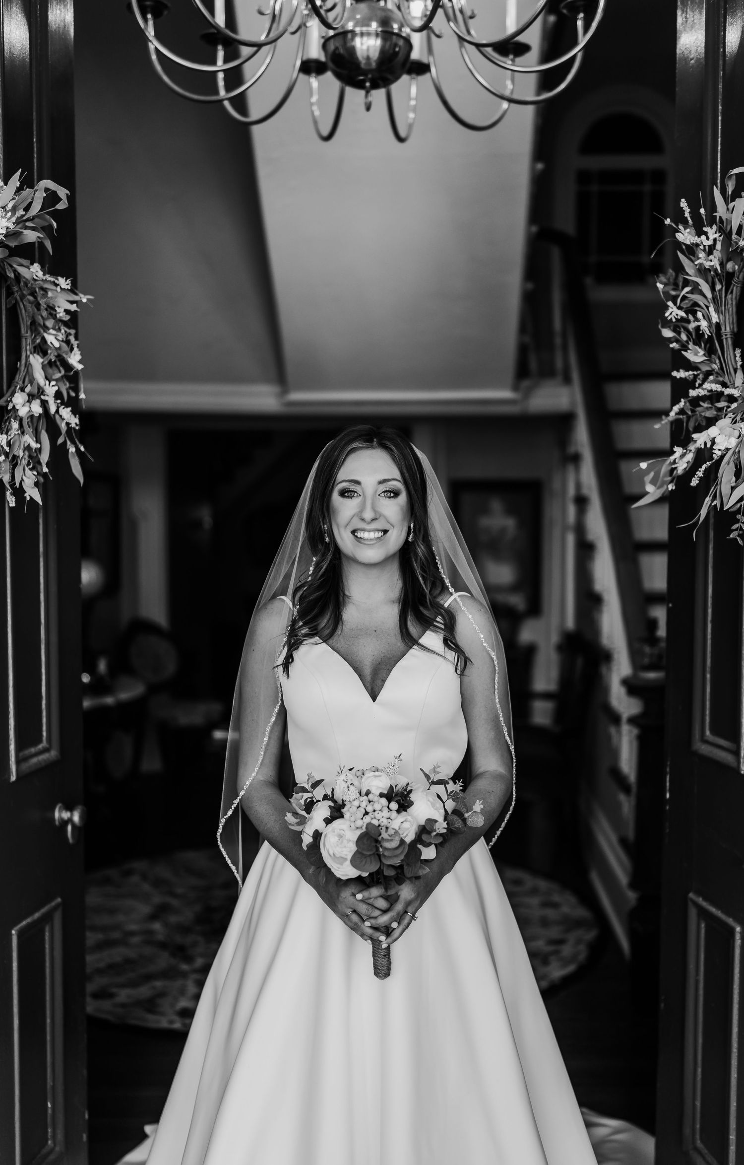Black and white portrait of a bride in a sweeping gown standing beneath an ornate chandelier.