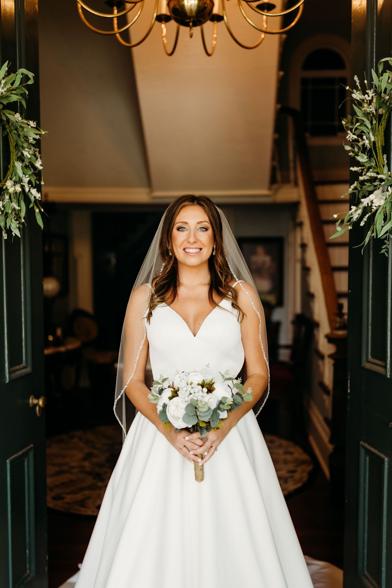 Bride in a stunning white wedding dress poses with her bouquet in a beautifully lit doorway.