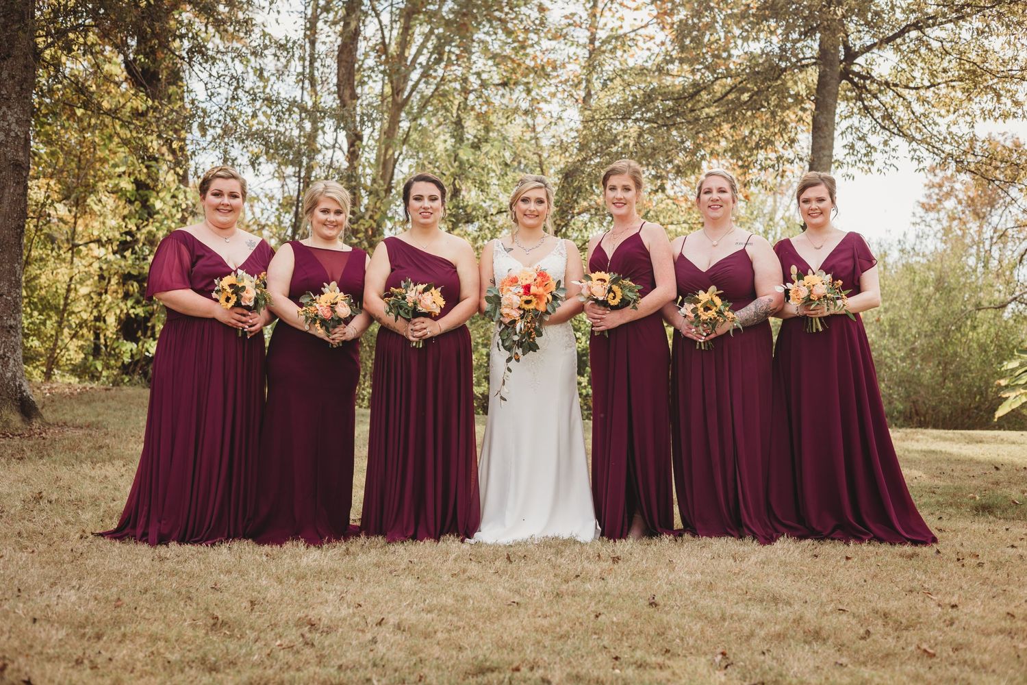 Wedding party in burgundy dresses standing in line against a fall foliage backdrop.