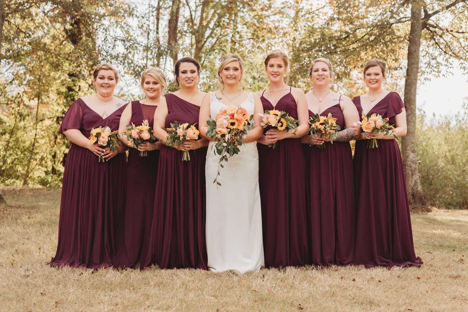 Outdoor wedding party portrait with bridesmaids in wine-colored dresses holding floral bouquets.