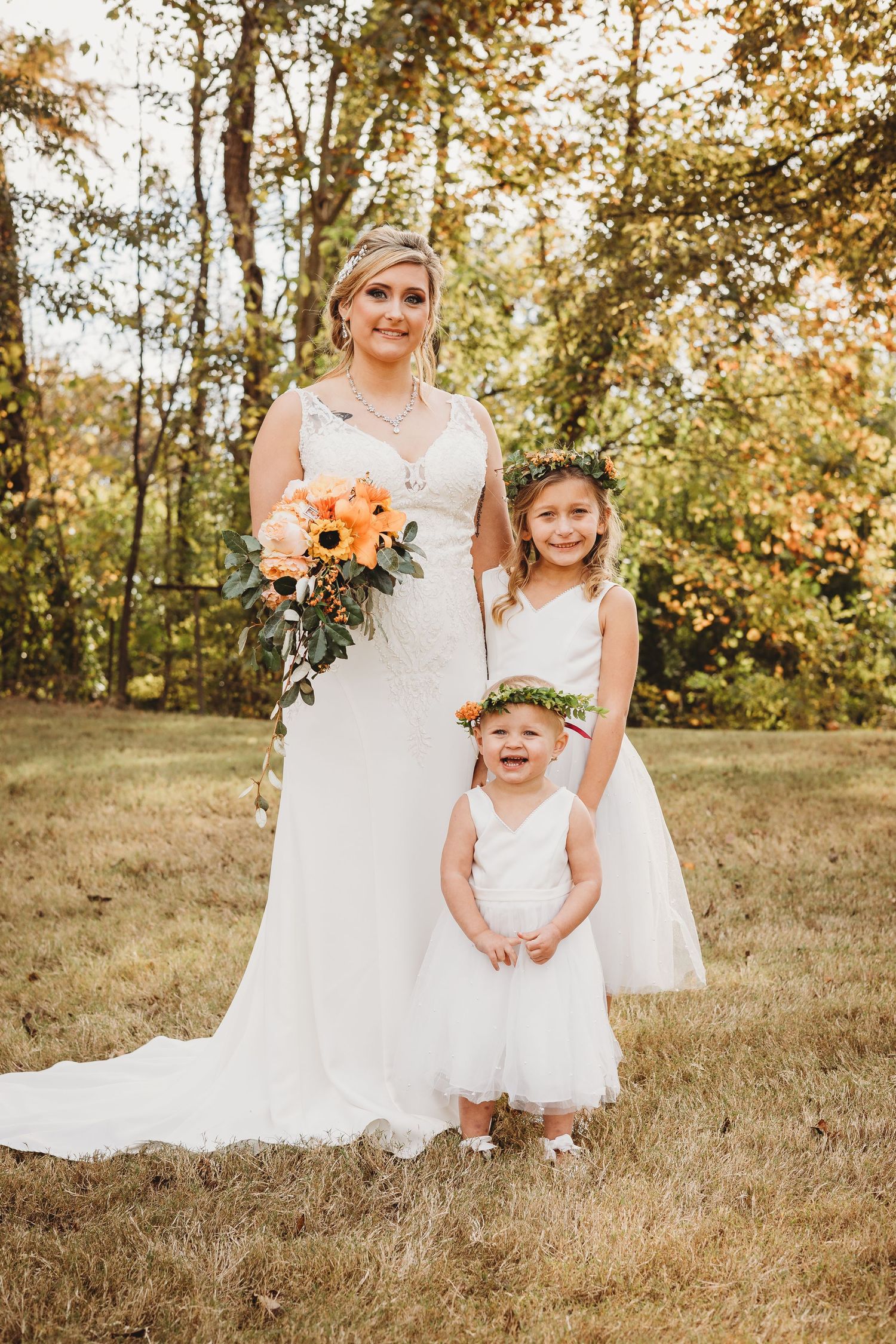 Wedding party members in white dresses pose with young flower girl in outdoor autumn setting.