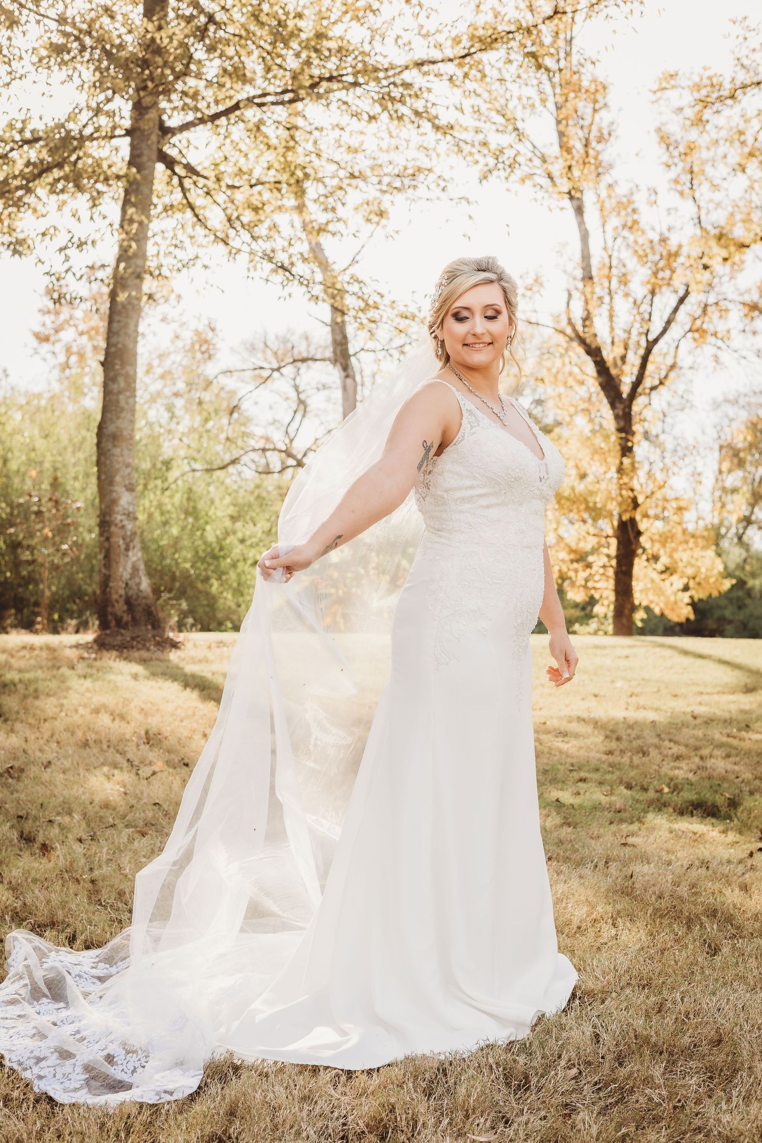 Bride in flowing white gown poses on grassy hillside during sunset.
