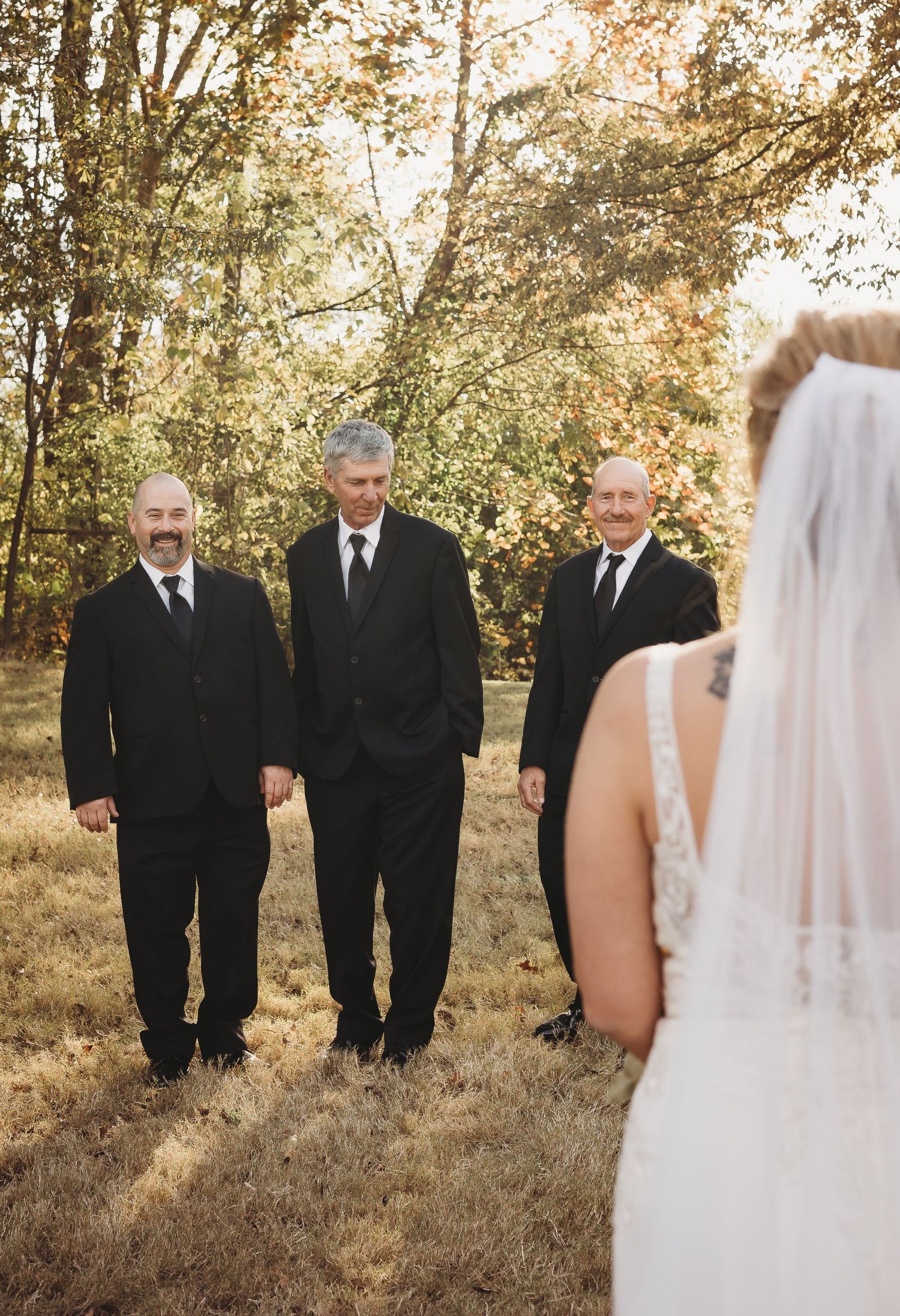 Men in black suits stand together in a grassy field during an autumn wedding photoshoot.