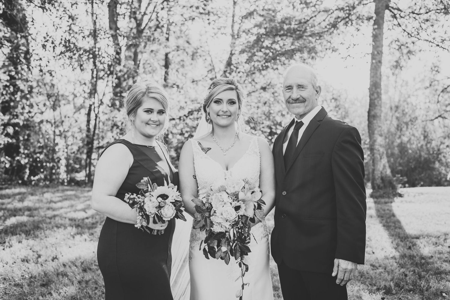 Wedding group stands together in black and white photograph with trees in background.