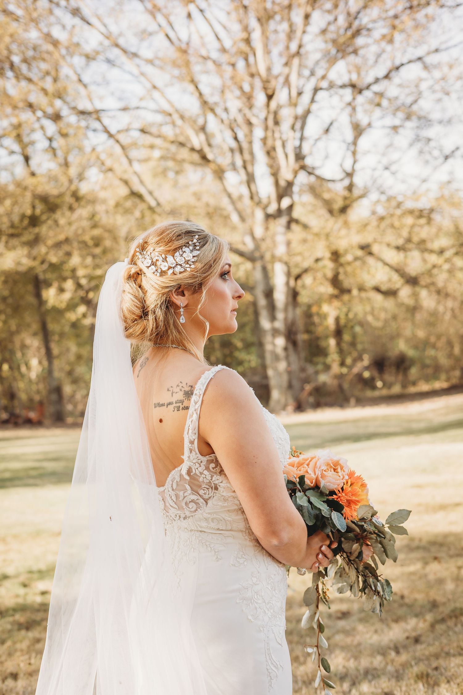 Bride in lace wedding dress with floral bouquet poses in autumn woodland setting during golden hour.