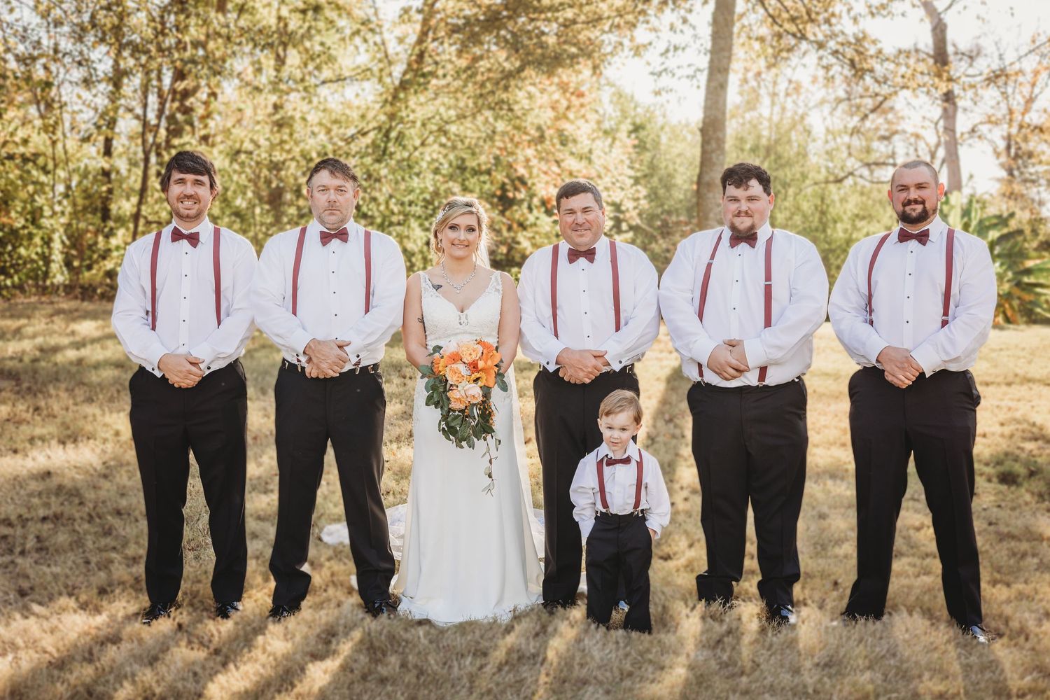 Wedding party members in black suits and red suspenders pose together in an autumn outdoor setting.