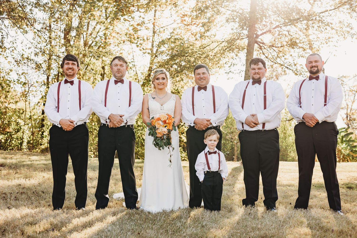 Wedding party members stand together in formal attire against a sunlit autumn backdrop.