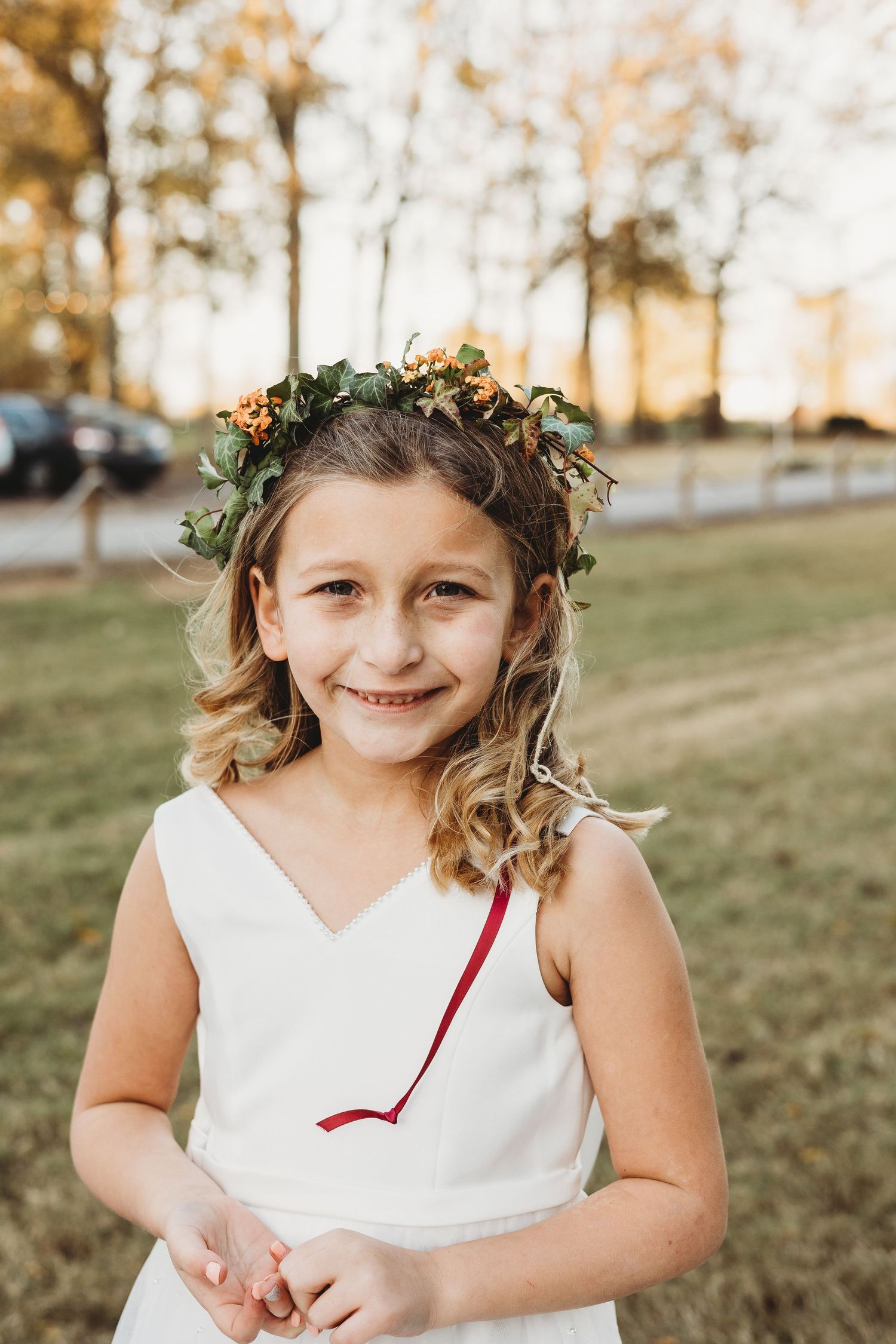 Young flower girl wearing white dress and floral crown smiles during outdoor wedding photos.