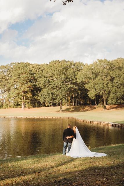 A couple in wedding attire poses by a serene lake surrounded by trees during golden hour.