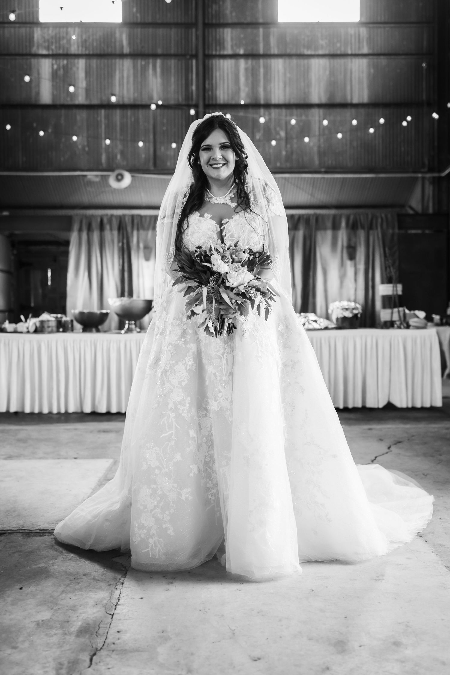 Black and white photo of bride in elegant lace gown with train in rustic venue.