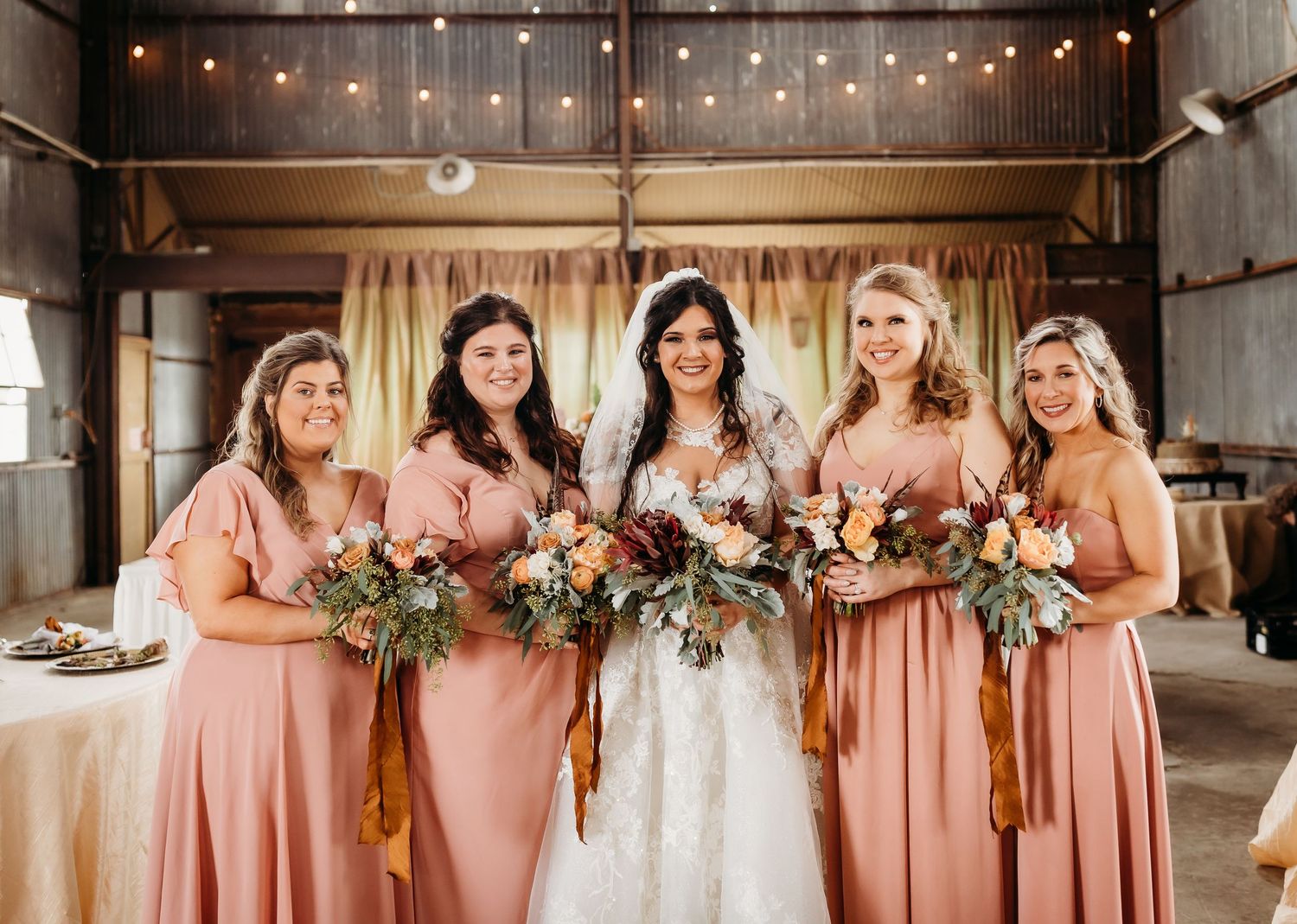 Wedding party in peach bridesmaid dresses poses with bride in rustic barn setting.