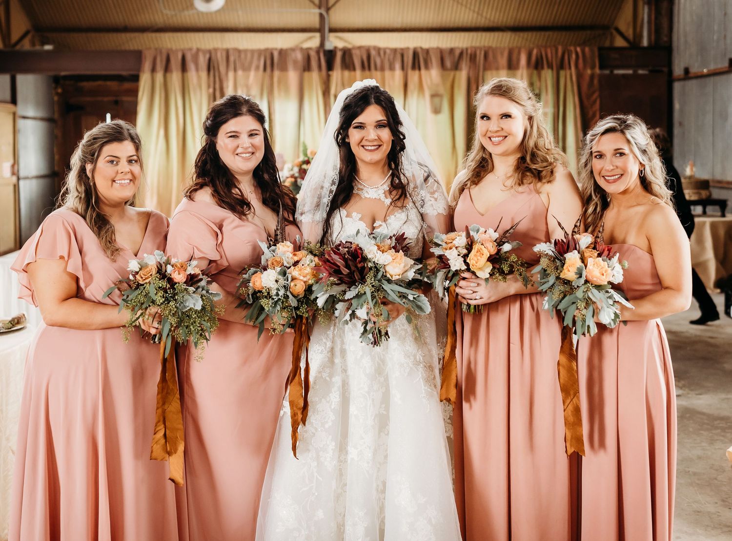 Bridal party wearing peach dresses stands in line with bride in white gown inside rustic venue.