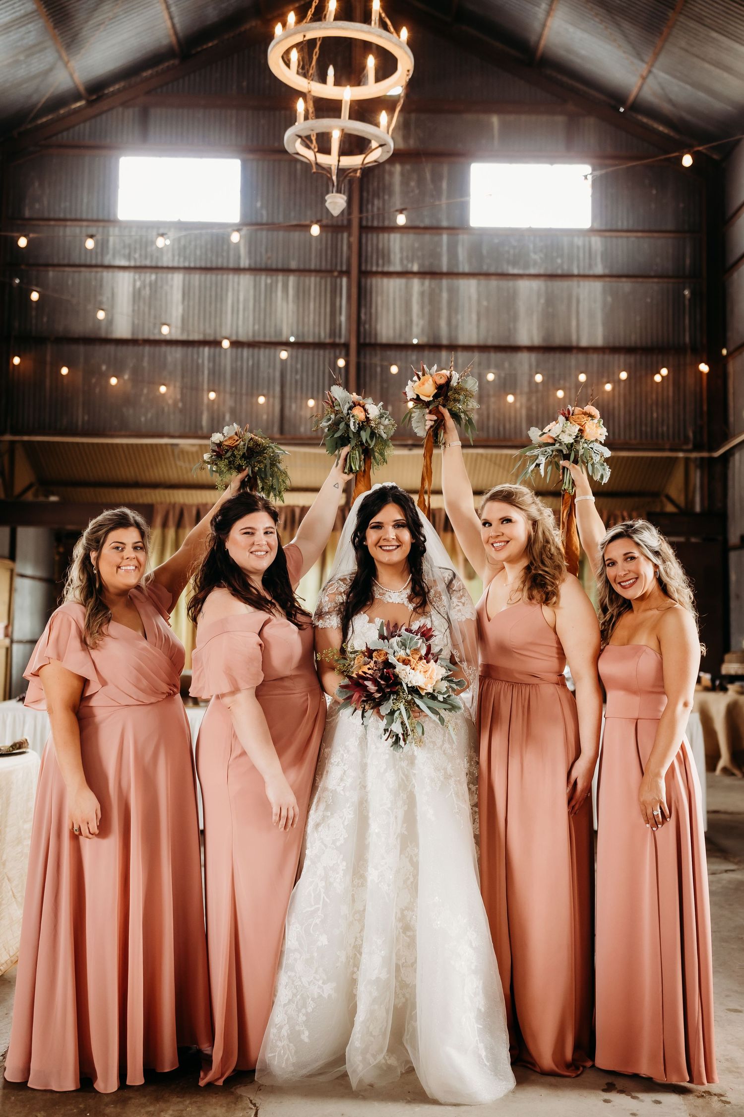 Wedding party in peach dresses celebrates with bride under rustic barn chandeliers.