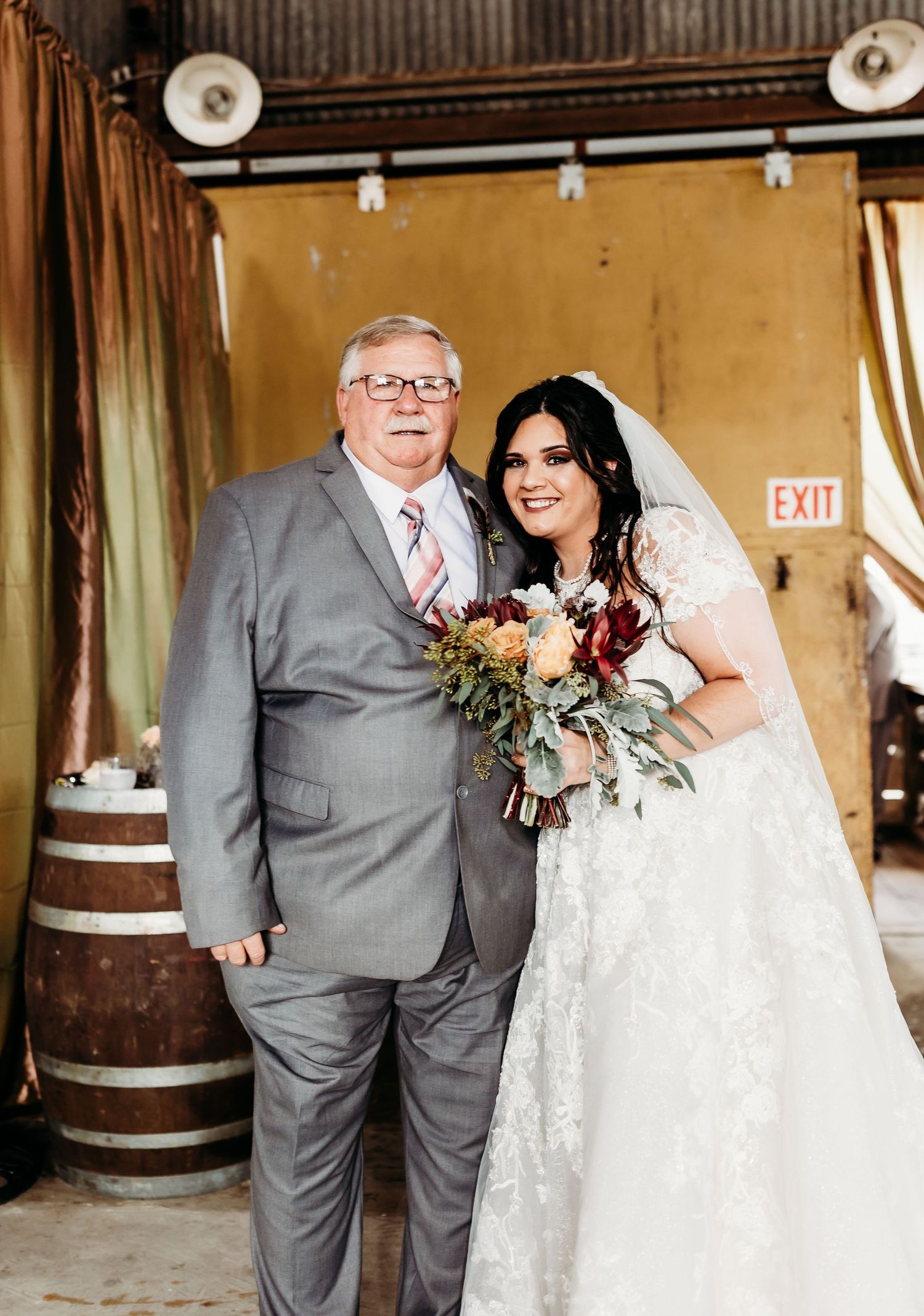Wedding party poses in rustic barn venue with wooden barrels and string light decorations.
