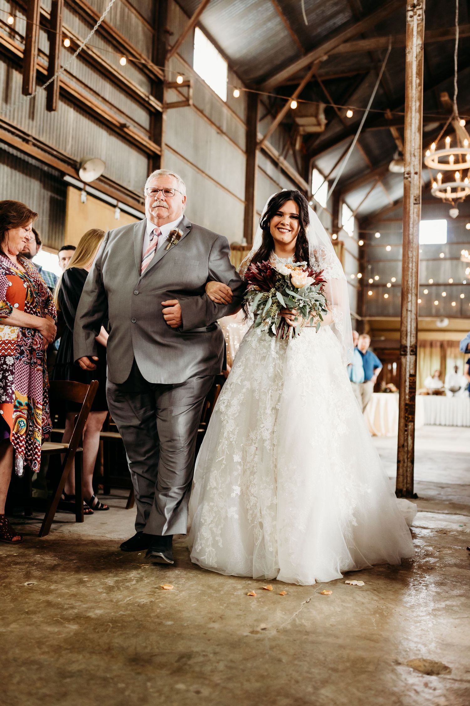 Wedding party walks down the aisle in an industrial venue with string lights overhead.