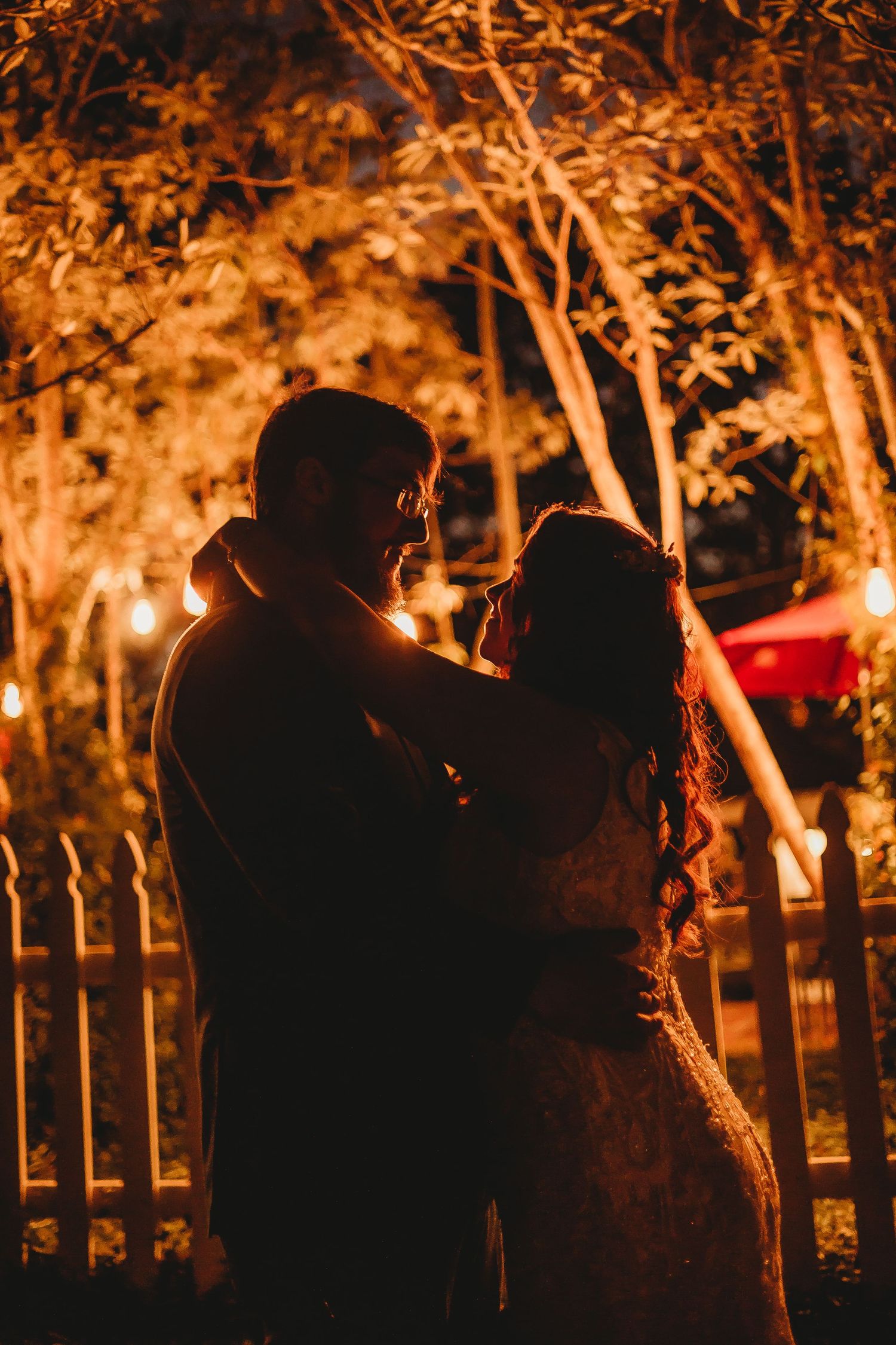 Silhouettes of wedding guests under illuminated trees at night with warm lighting.