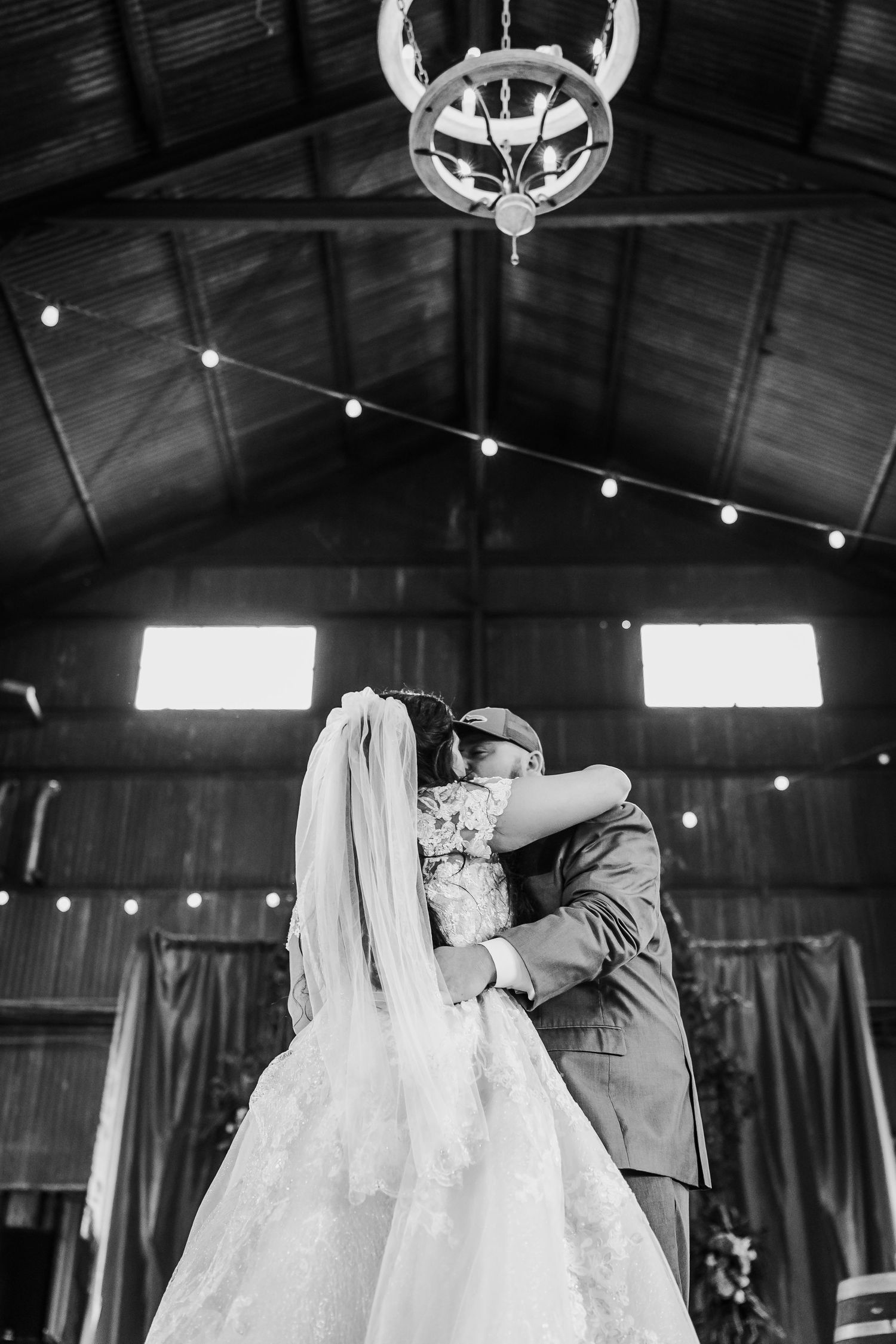 Black and white photo of newlyweds dancing in rustic venue with string lights.