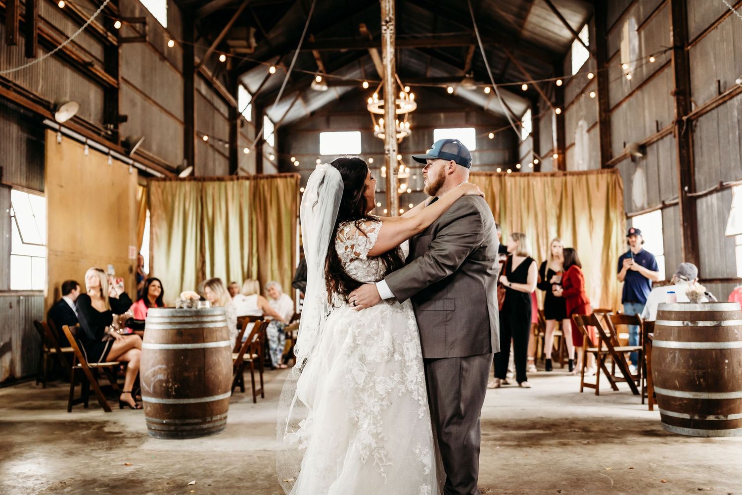 A couple dances together in a rustic barn venue decorated with string lights and wooden barrels.