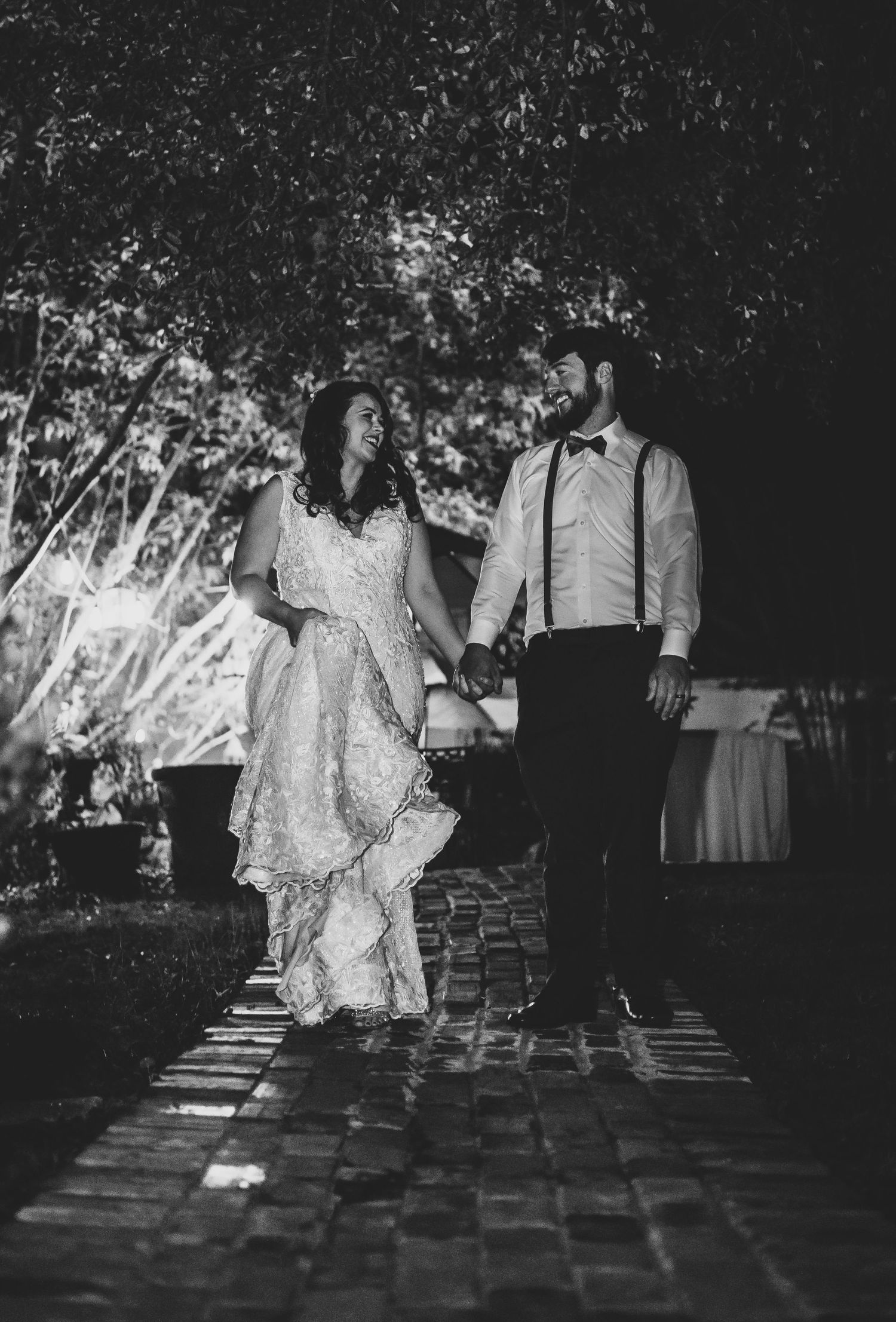 Black and white photo of wedding party walking on cobblestone path under trees.