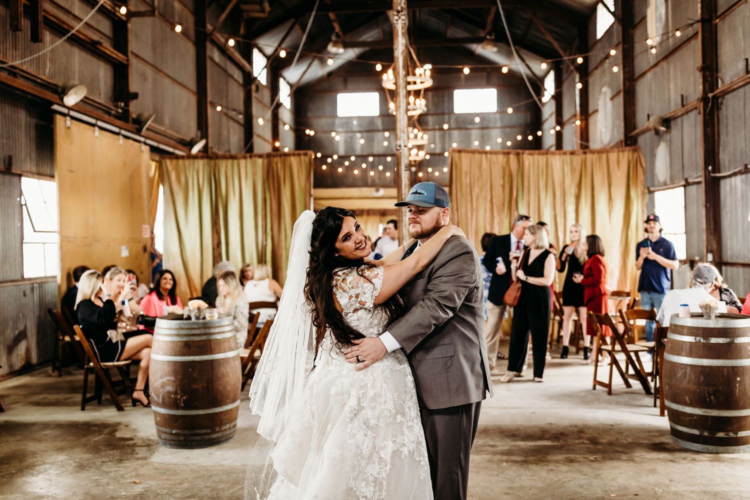 Newlyweds dance in a rustic barn venue with string lights and wooden barrel decor.